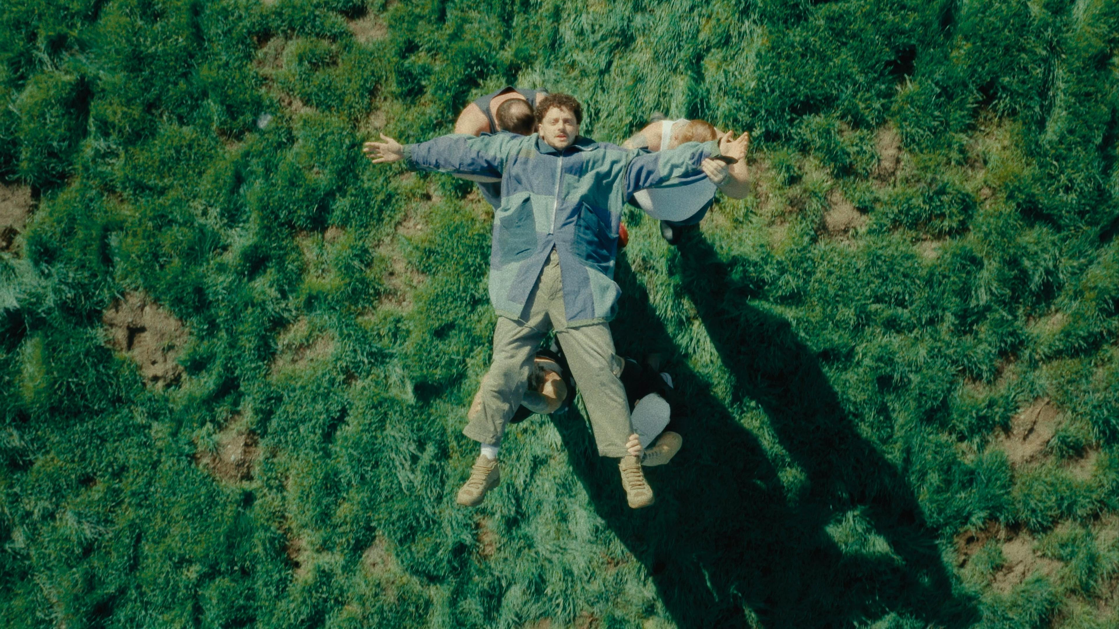 Thumbnail for Grandson's music video 'Eulogy' depicting the artist lying flat on his back, arms and legs outstretched, as he is being carried by four people across a field of lush green grass. Grandson is dressed in a pale blue jacket and beige pants, contrasting with the natural greenery below him.