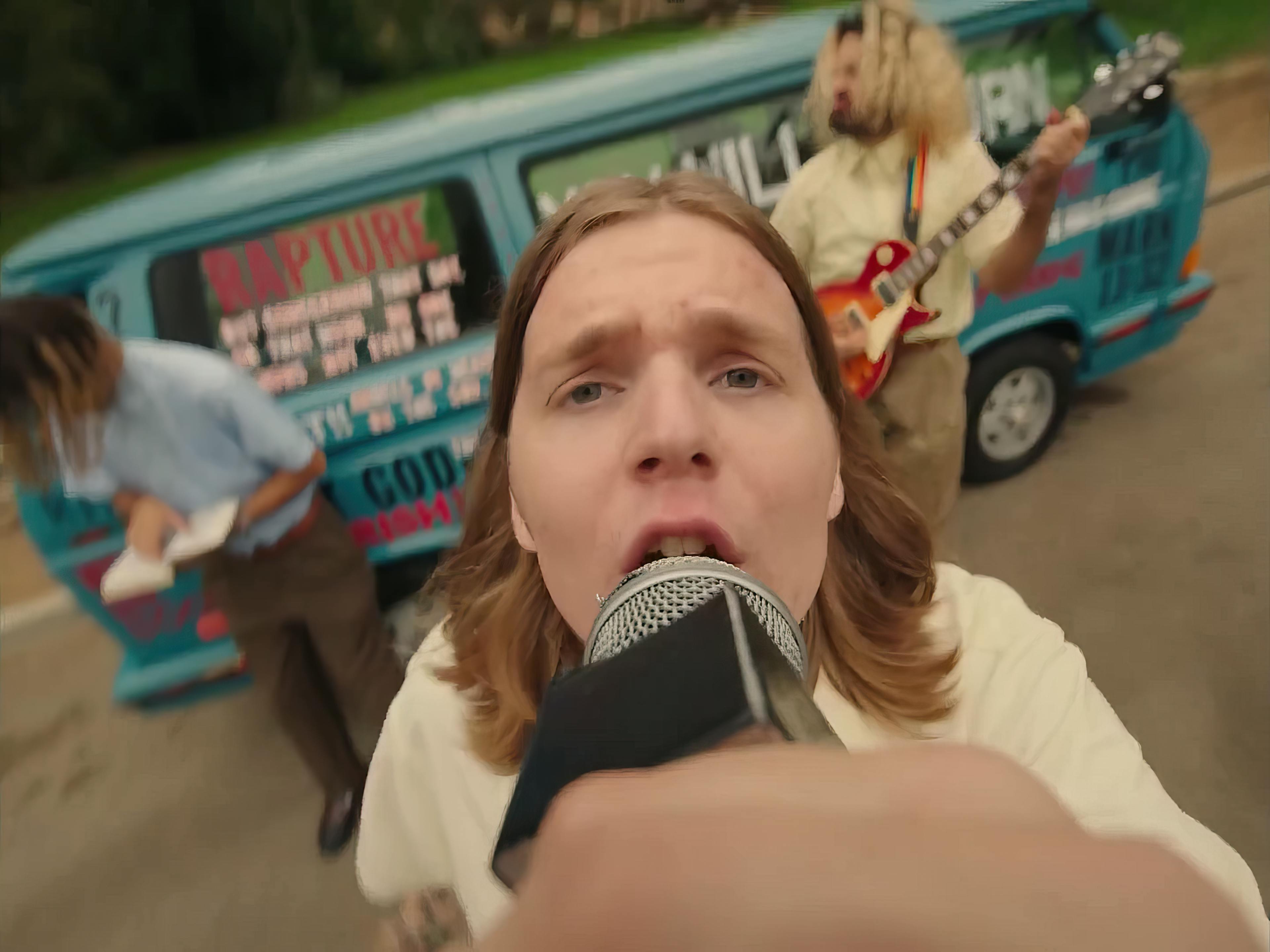 The lead singer of JAWNY with shoulder-length hair sings passionately into a microphone. Behind him, out of focus, are band members and a van detailed with vibrant text and graphics suggesting a dynamic music event. The setting seems informal and energetic, typical of a music video setting.