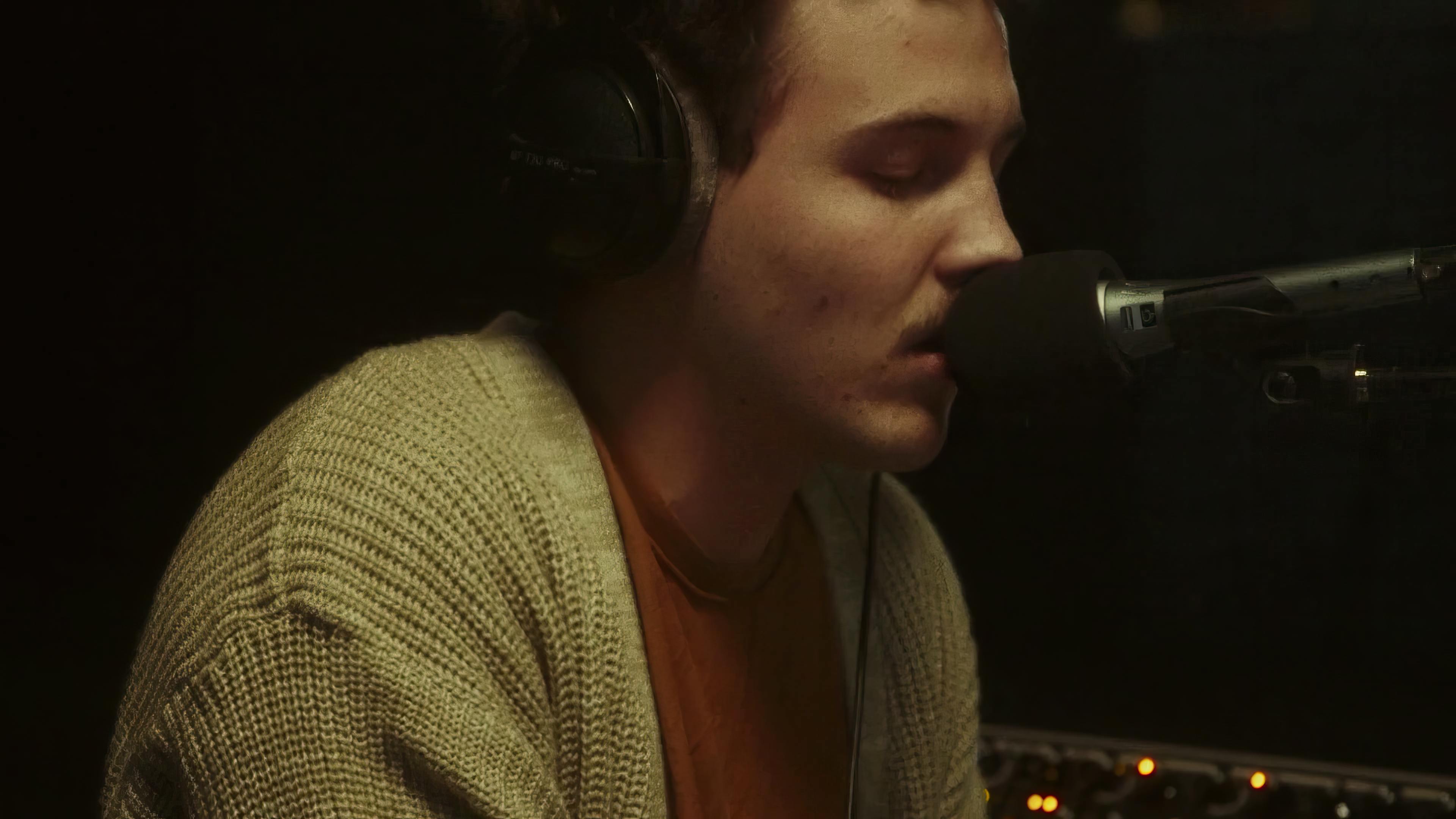 In a dimly lit room, an artist, Ritt Momney, is captured mid-performance during a live session. He's engrossed in singing into a microphone, wearing headphones and a cozy beige cardigan over an orange shirt. His closed eyes and serene expression suggest a deep connection to the music he's creating, conveying a sense of intimacy and focus in the moment.