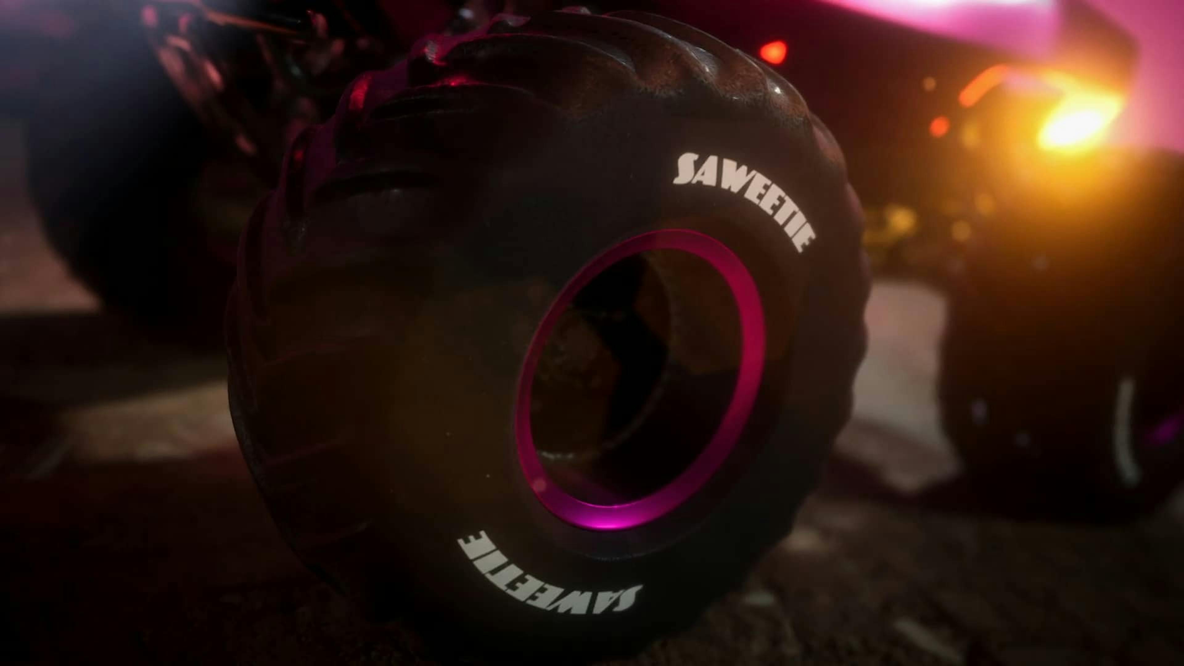 A dramatic close-up of a large, black off-road tire with the name 'SAWEETIE' printed in bold white letters on its side. The inner rim of the tire features a vivid pink glow, giving the image a dynamic and edgy feel. The background is dimly lit, with a hint of the vehicle's body and ambient lights that suggest a powerful, rugged aesthetic associated with high-energy action.