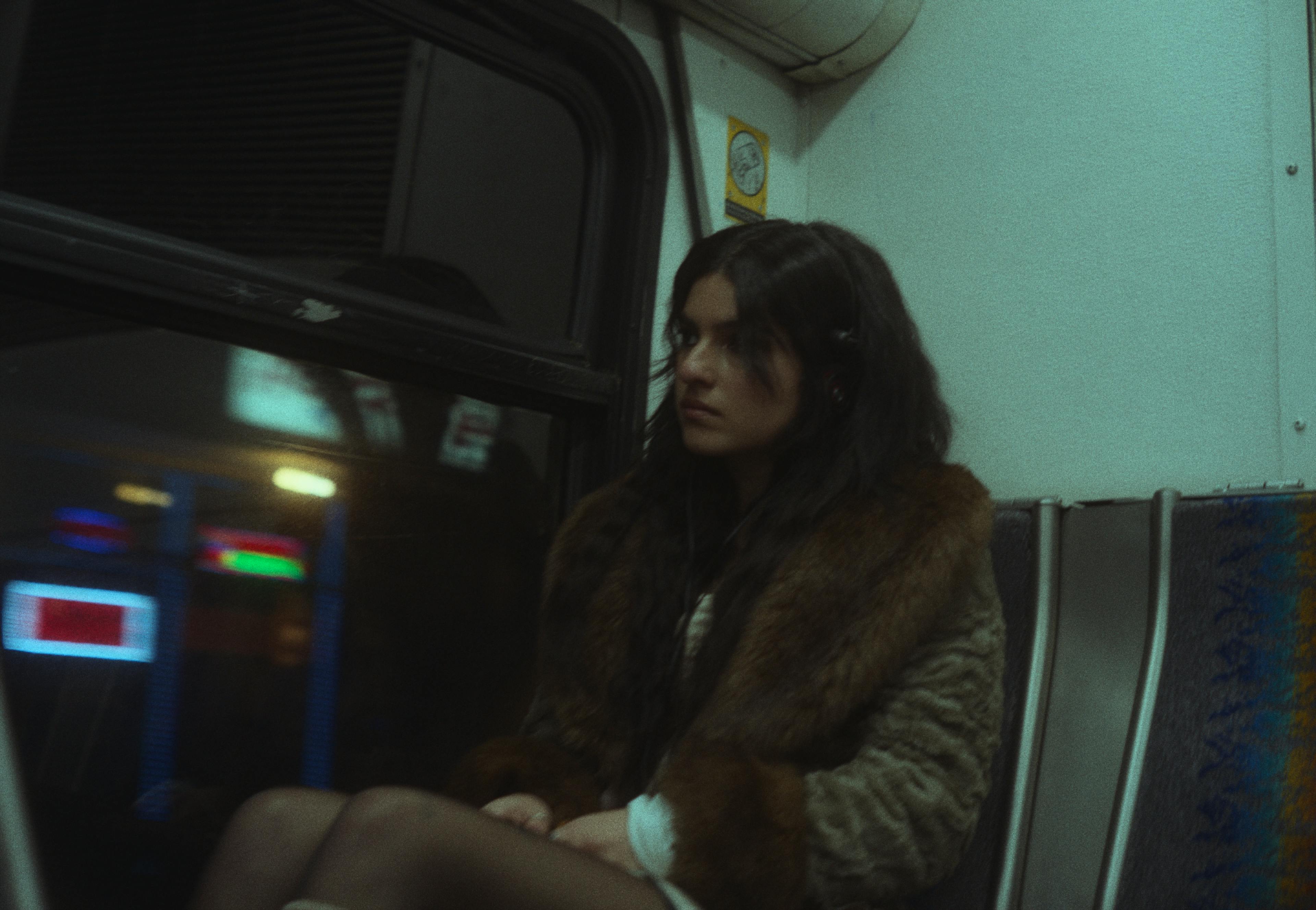 A young woman, Ren, seated inside a bus at night. She is wearing a large, fluffy brown faux fur coat, and her long, dark hair is partly obscuring her face.