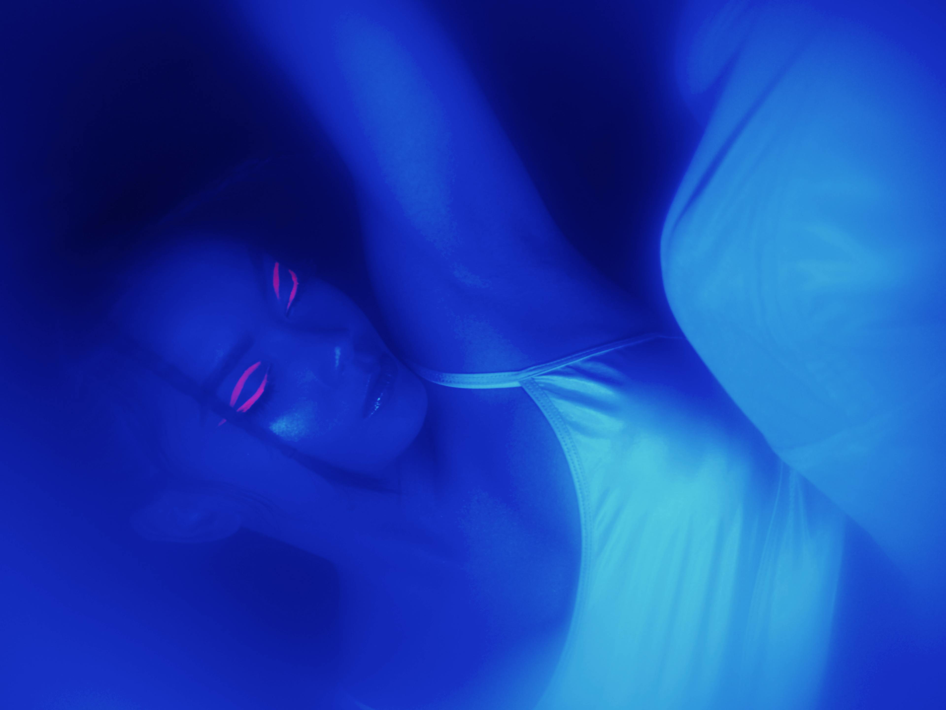 Ylona Garcia sleeps in a blue incubation chamber with glow in the dark pink eyeliner and a white croptop.