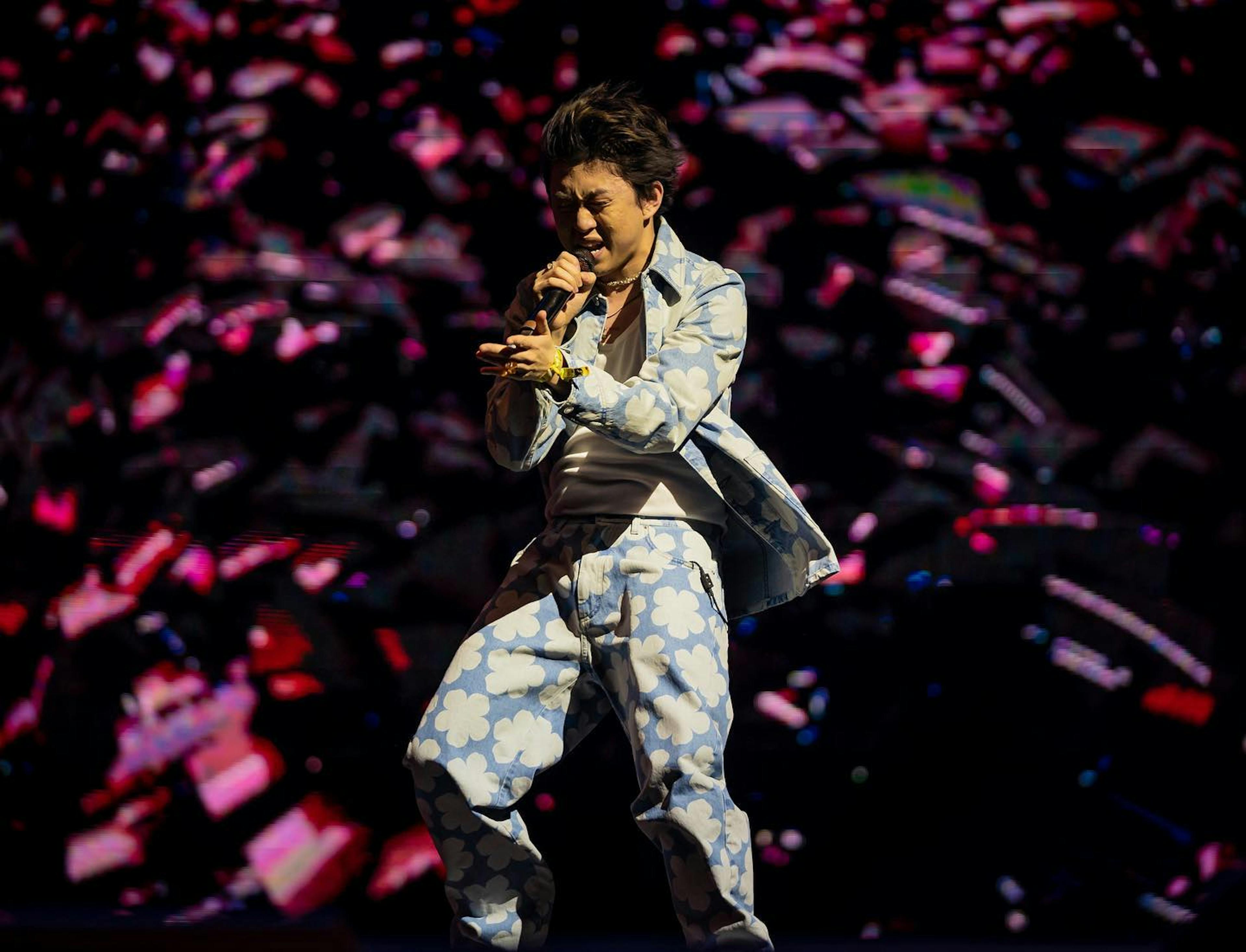 Rich Brian is performing energetically on stage at Coachella. He's donning a light blue and white floral suit, infusing his dynamic stage presence with style. With his hair swept back and eyes closed, he sings into the microphone with intense emotion. The backdrop is a riot of abstract pink and blue lights, casting a vibrant glow that captures the festival's electric atmosphere and the artist's passionate performance.
