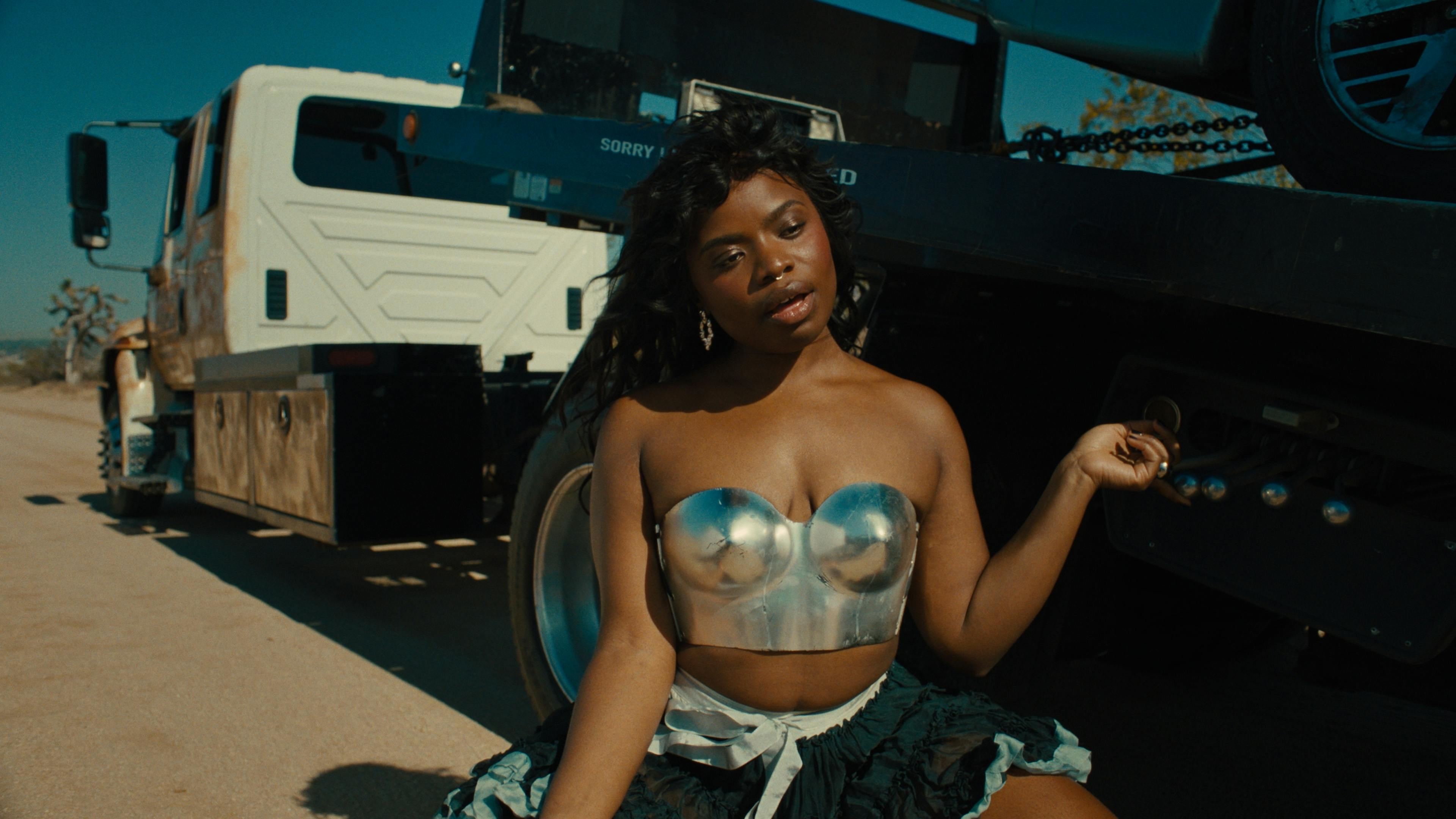Vagabon sits in front of a large truck. She's wearing a metallic silver bralette and a ruffled black skirt, with a natural makeup look and her hair styled in loose waves. The setting is outdoors on a sunny day, with clear skies and a dry landscape in the background.