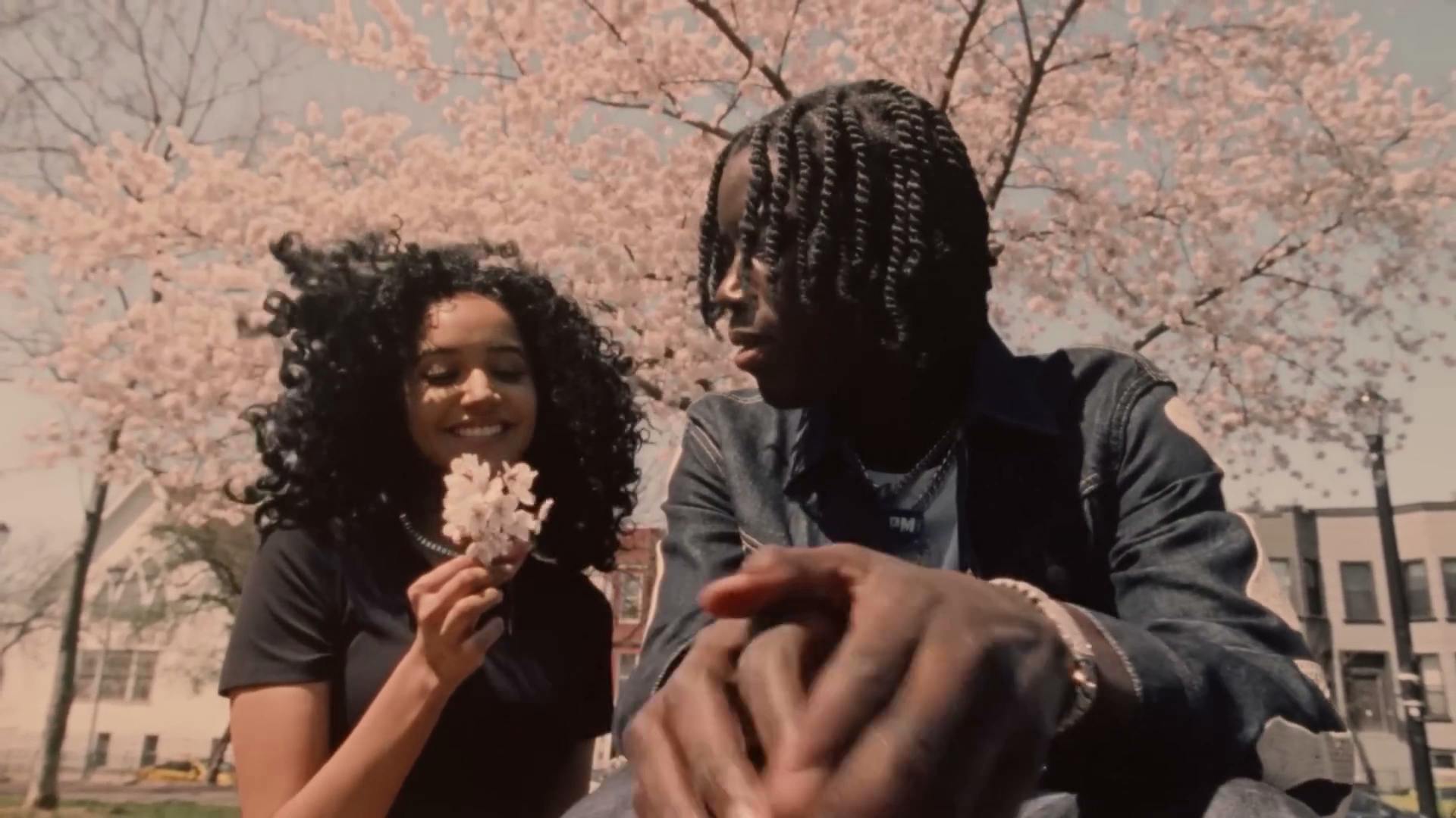 A woman and Melvoni sit side by side under a cherry blossom tree, the woman smiling and holding a sprig of pink blossoms. Melvoni, dressed in a denim jacket, looks at her. They are surrounded by the soft hues of spring and a clear sky.