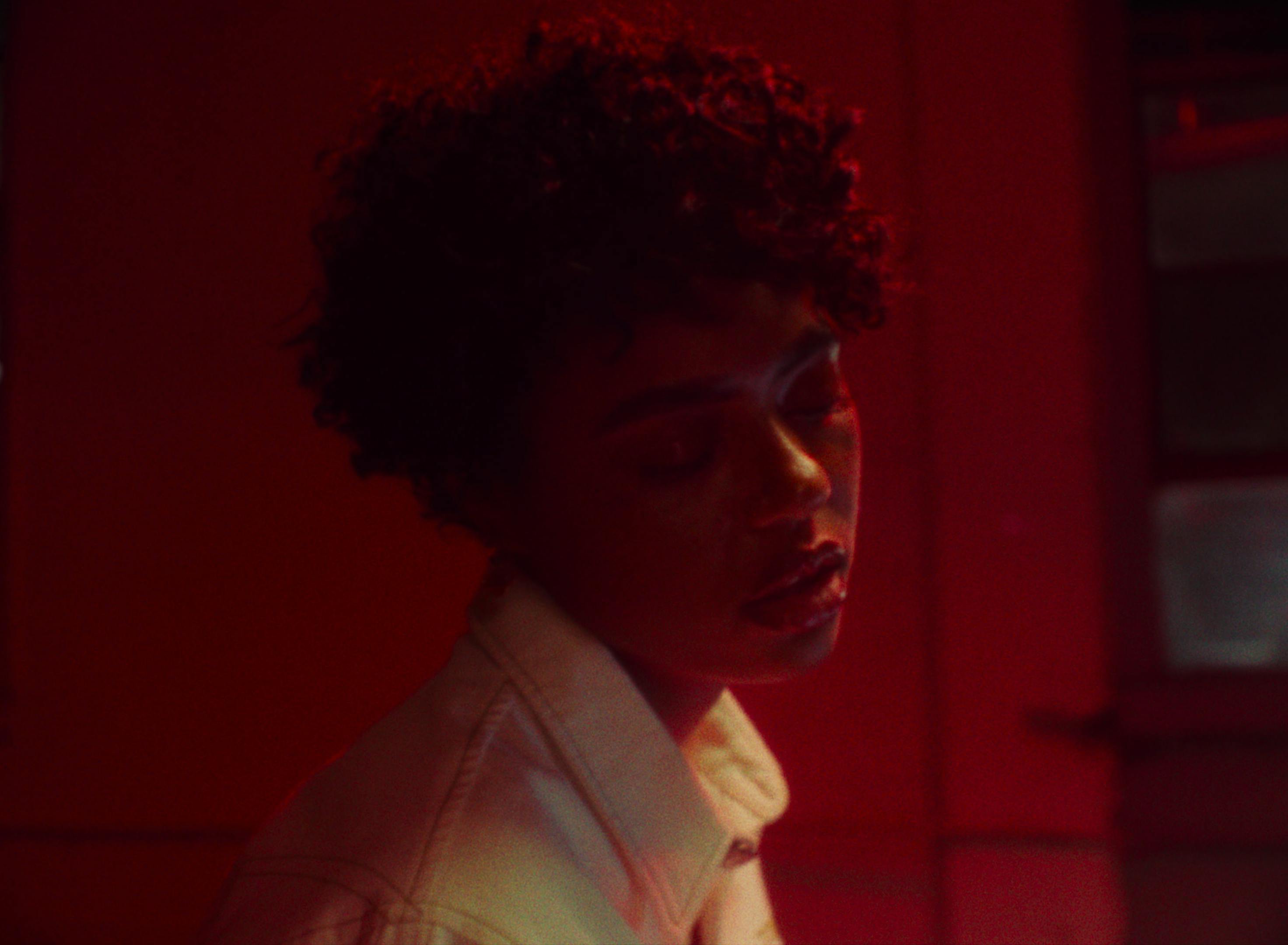 A still from Samaria's music video 'Out The Way' depicts a woman in a contemplative state. The scene is bathed in a red hue, casting dramatic shadows on her face and highlighting her short curly hair. She's wearing a white jacket, and her profile is captured against a darker, blurred background, adding a sense of depth and introspection to the image. Her expression is somber and thoughtful, resonating with the emotional tone of the song.