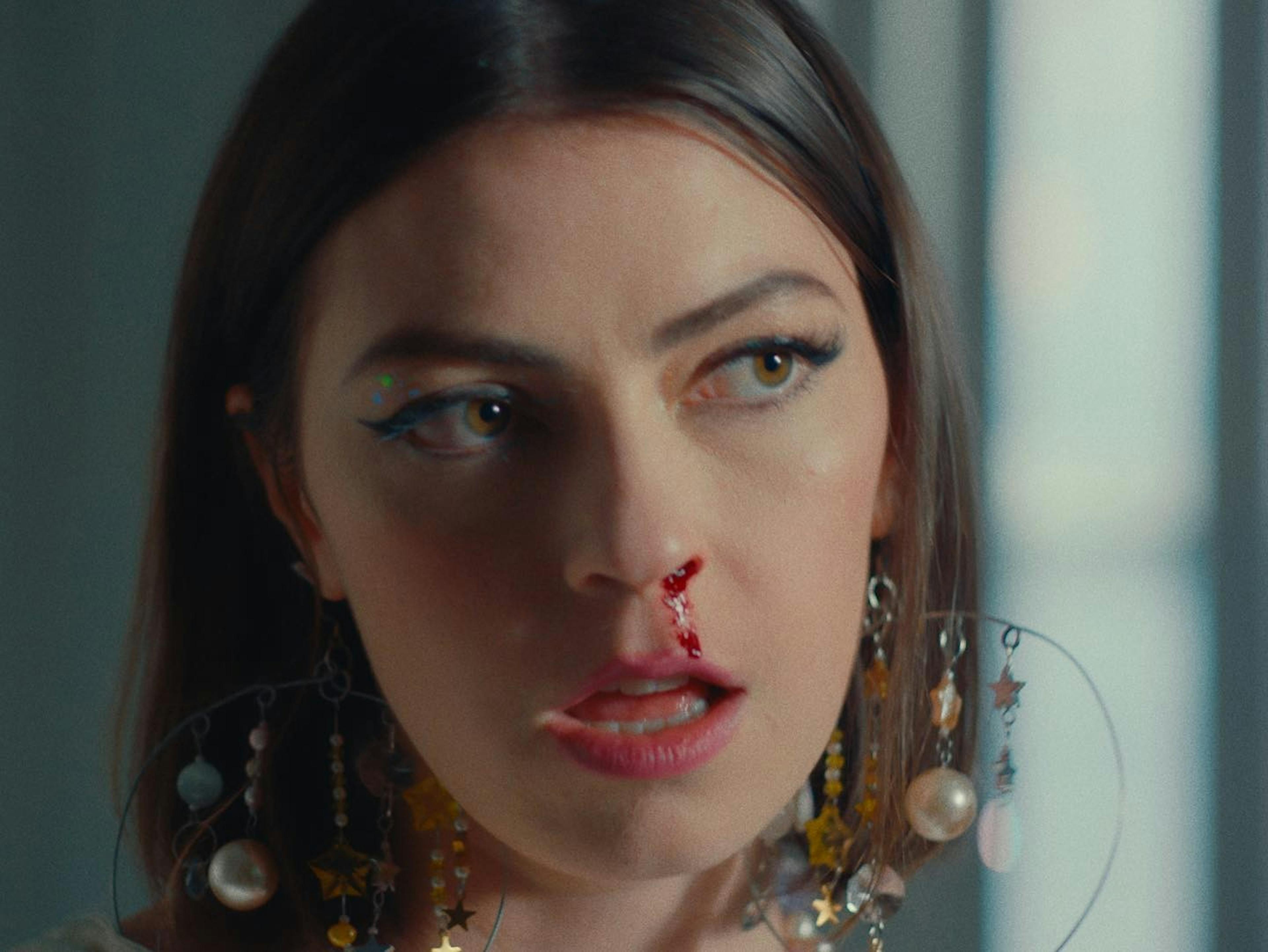A close-up still from Anna Shoemaker's music video 'Everything Is Fine' shows the artist with a pensive expression. She has distinctive makeup with a blue winged eyeliner and a colorful dot near the corner of her eye. Large, ornate earrings with stars and pearls adorn her ears. There's a trickle of blood coming from her nostril, adding a dramatic and possibly symbolic element to the scene. Natural light from a window casts soft illumination on her face, highlighting her features against a neutral background.
