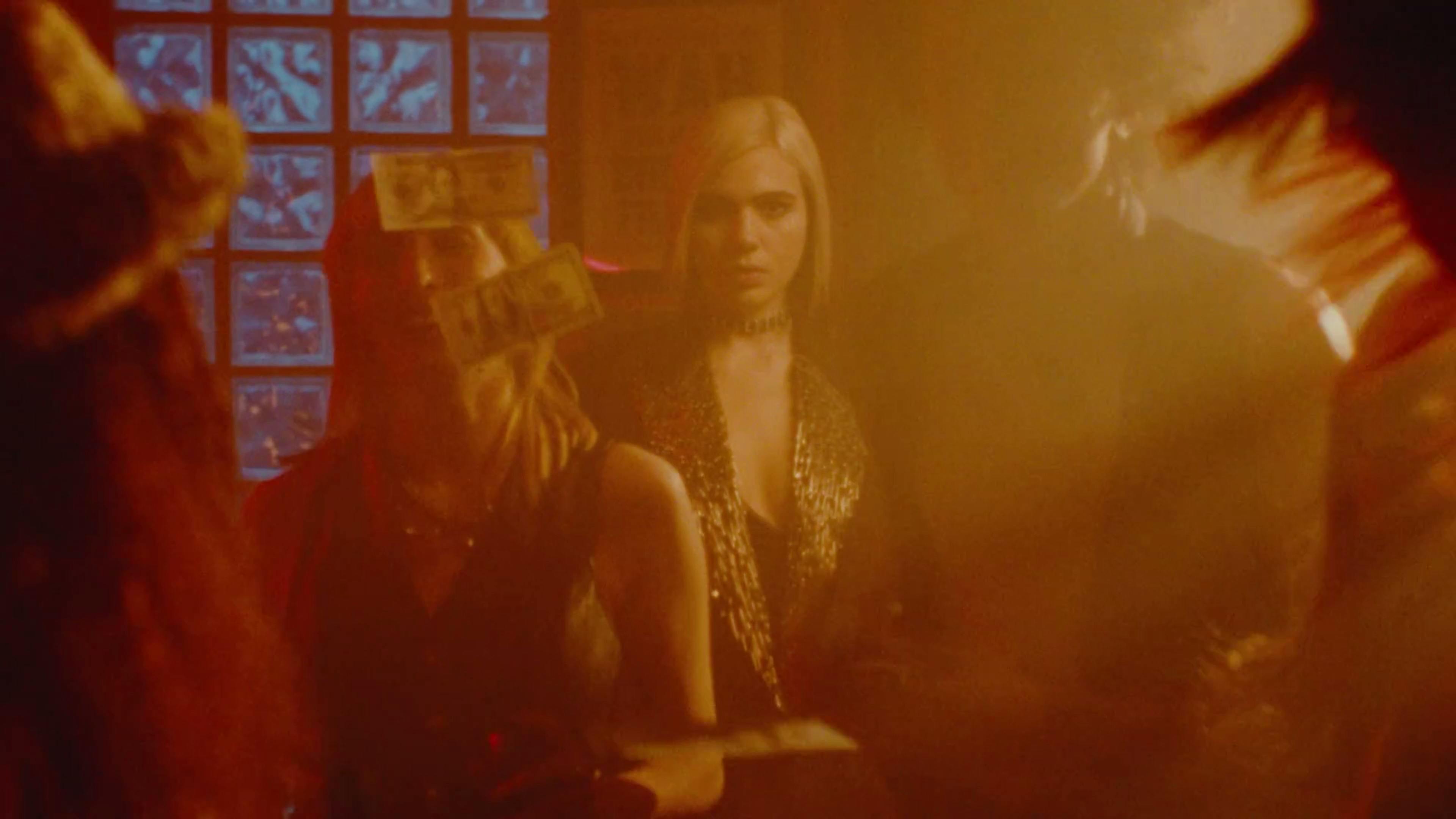 A hazy, reddish-toned still from Sunflower Bean's music video 'Roll The Dice' shows a woman with a stern expression, gazing directly at the camera. She's adorned with a glittering, chain-like garment, and the blurred foreground suggests movement. The soft focus and warm lighting create a moody and mysterious atmosphere.