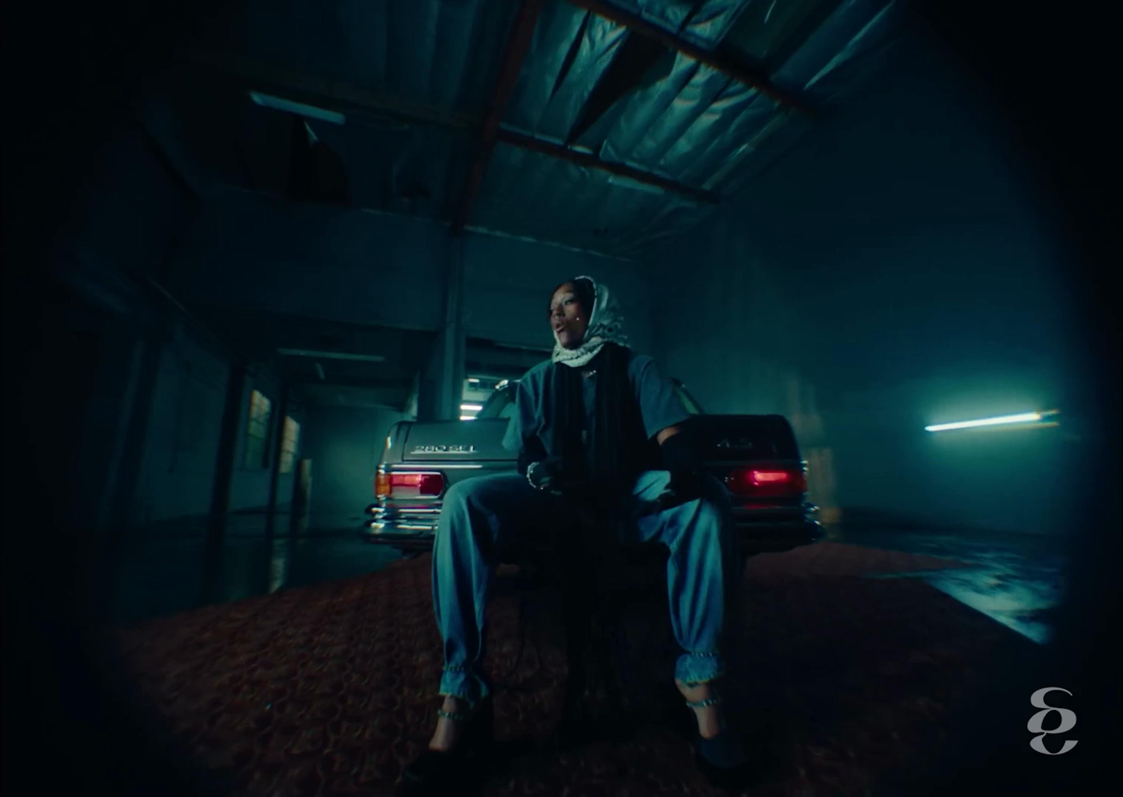 In a dimly lit garage setting from Aryèe The Gem's music video 'To It,' she sits confidently on a vintage car. She's wearing a grey hooded top, blue jeans, and black heels. The ambient lighting casts a blue tone across the scene, with streaks of light piercing the background, creating a dramatic and stylized visual.