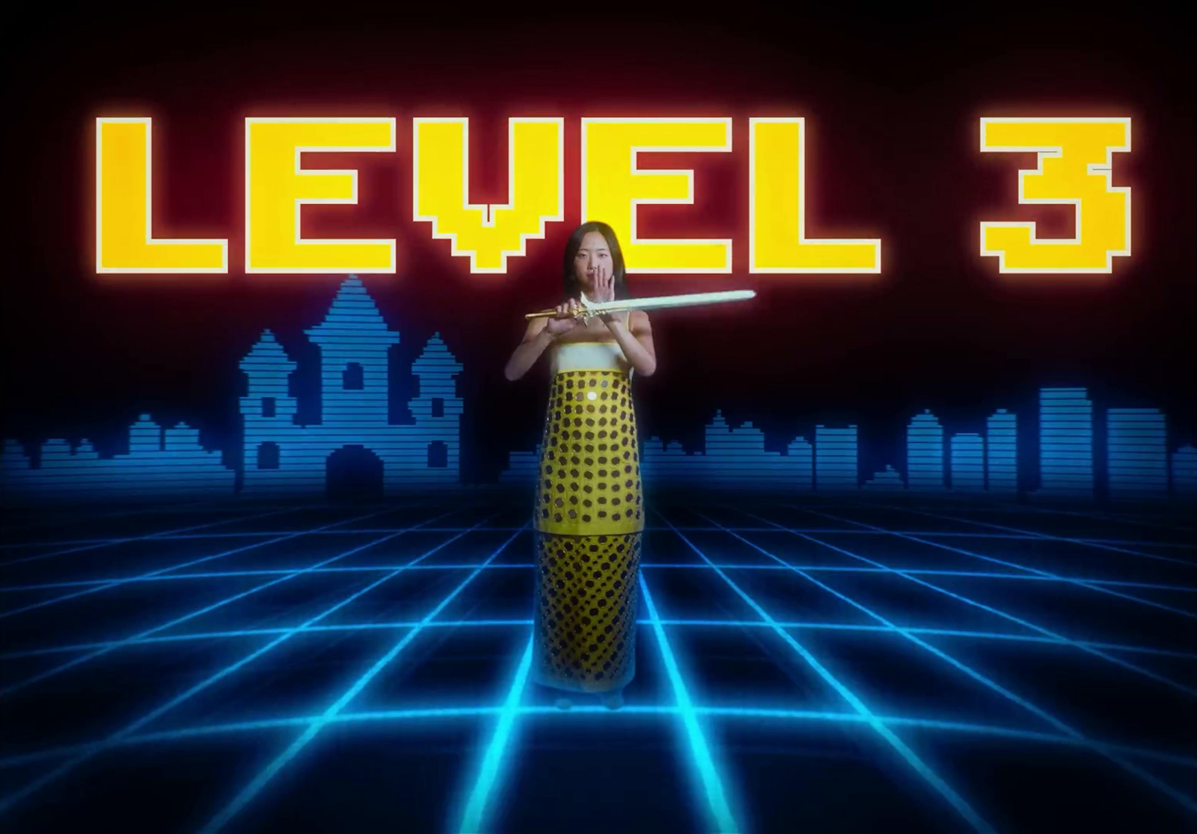 Sarah Kinsley stands centered in a vibrant, retro video game-inspired environment from her music video 'Oh No Darling!'. She's dressed in a traditional yellow garment with black polka dots, holding a sword horizontally in front of her. The backdrop features large, pixelated yellow letters spelling "LEVEL 3", with digital outlines of castles and a cityscape on a grid that glows with neon blue lines, reminiscent of a classic arcade game setting.