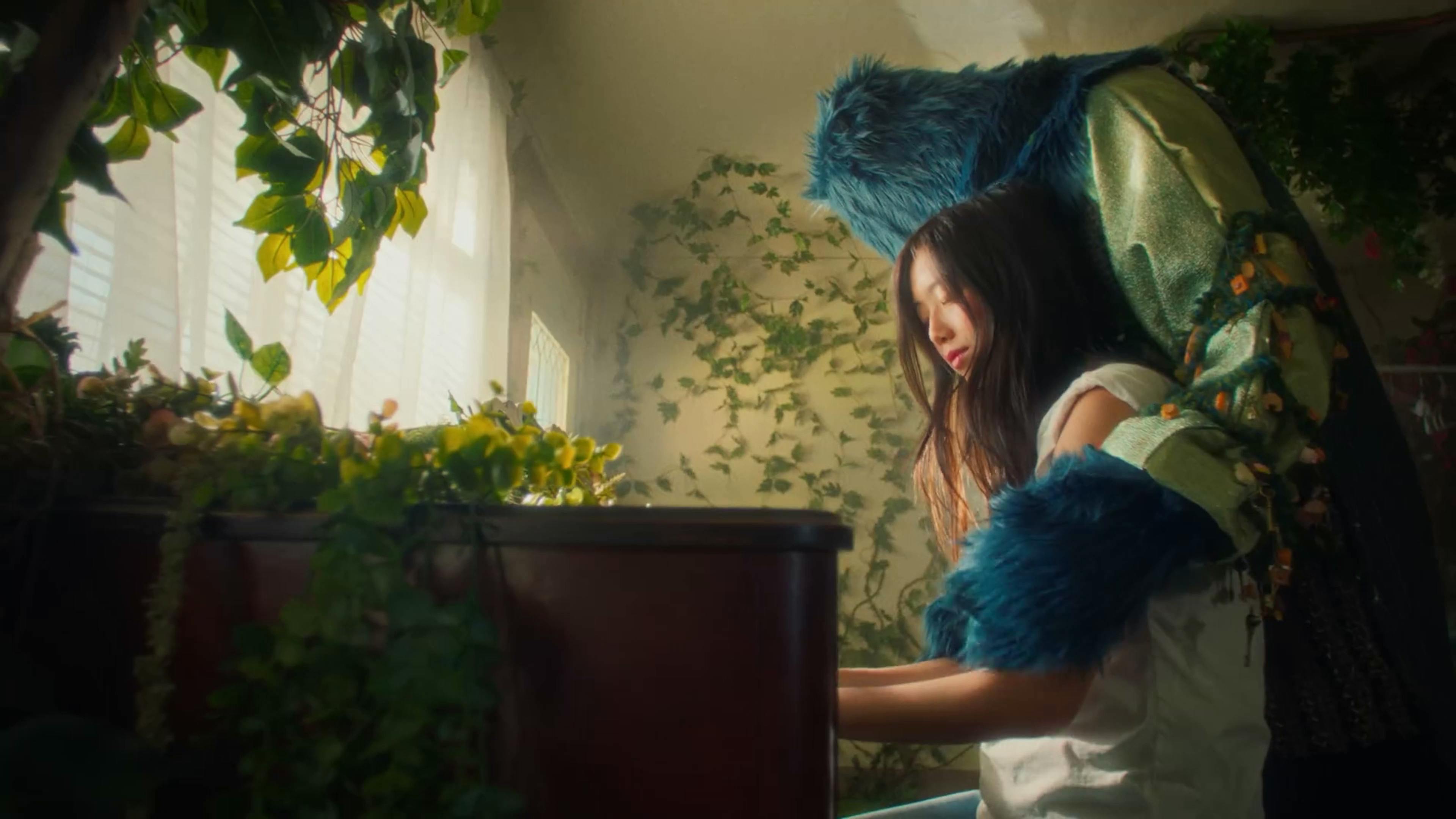 A whimsical still from Sarah Kinsley's music video 'Lovegod' portrays the artist and a creature with blue fur and green embellishments in a sunlit room filled with plants. The artist appears serene as she tends to the greenery, while the creature, standing behind her, adds a touch of fantasy to the scene. The natural light streaming through the blinds creates a warm, inviting atmosphere.
