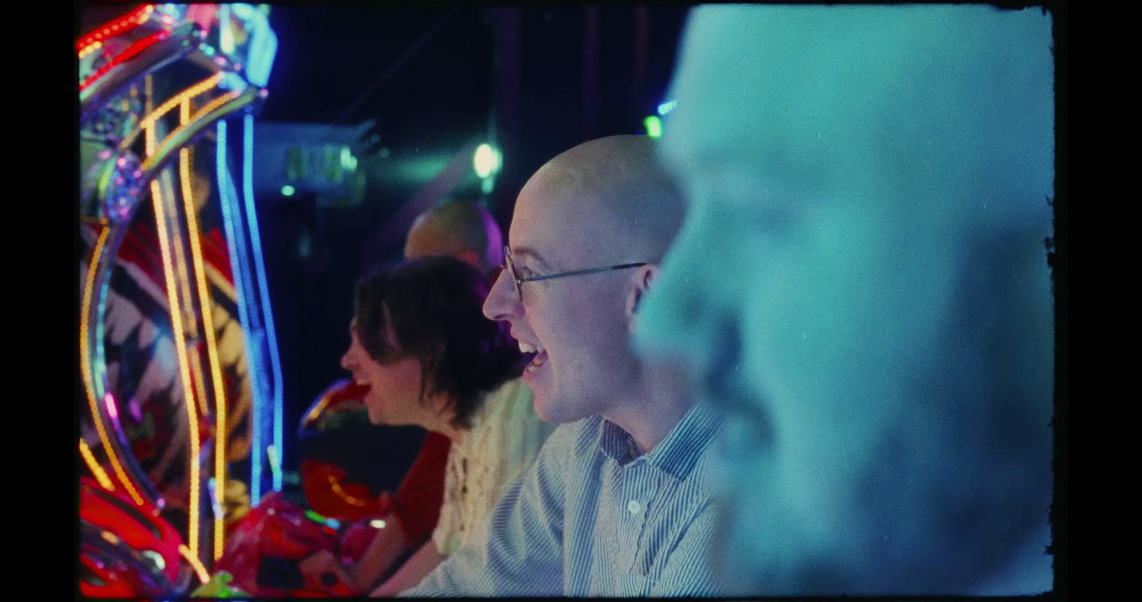 A still from Bombay Bicycle Club's music video 'Tekken 2' captures a moment of joy at an arcade. A person with a broad smile is playing a game, immersed in the experience. They're wearing glasses and a striped shirt, and the arcade's vibrant, colorful lights reflect in their glasses. The image conveys a sense of nostalgia and the simple pleasure of gaming in a lively, neon-lit setting.