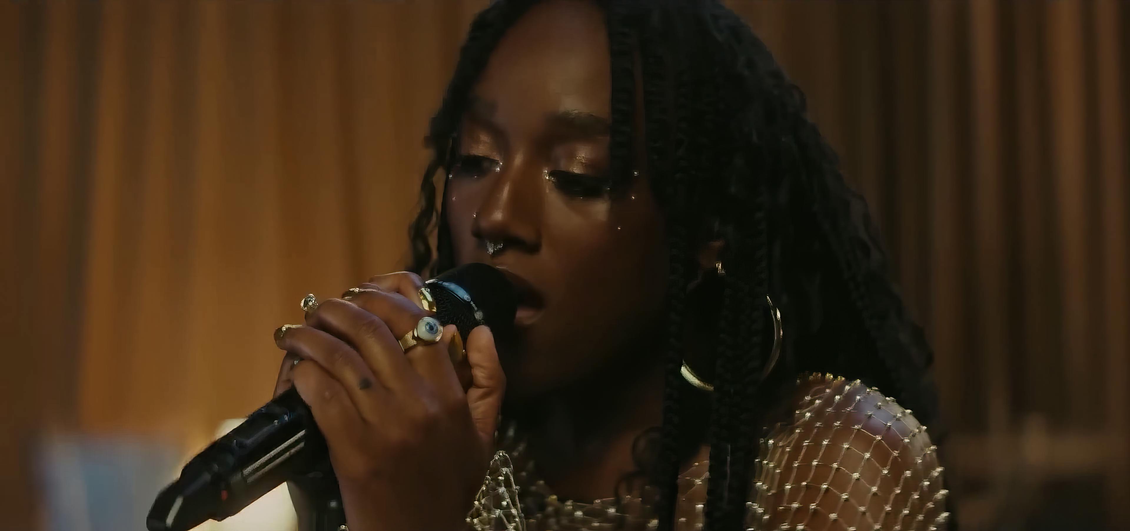 A close-up of Aryèe The Gem performing in a live session for the song 'Mirror.' She is holding a microphone close to her lips, eyes closed, conveying deep emotion. Her hair is styled in long braids, and she's adorned with shimmering eye makeup and sparkling earrings. The warm, ambient lighting and soft background lend a soulful atmosphere to the scene, focusing attention on her expressive performance.