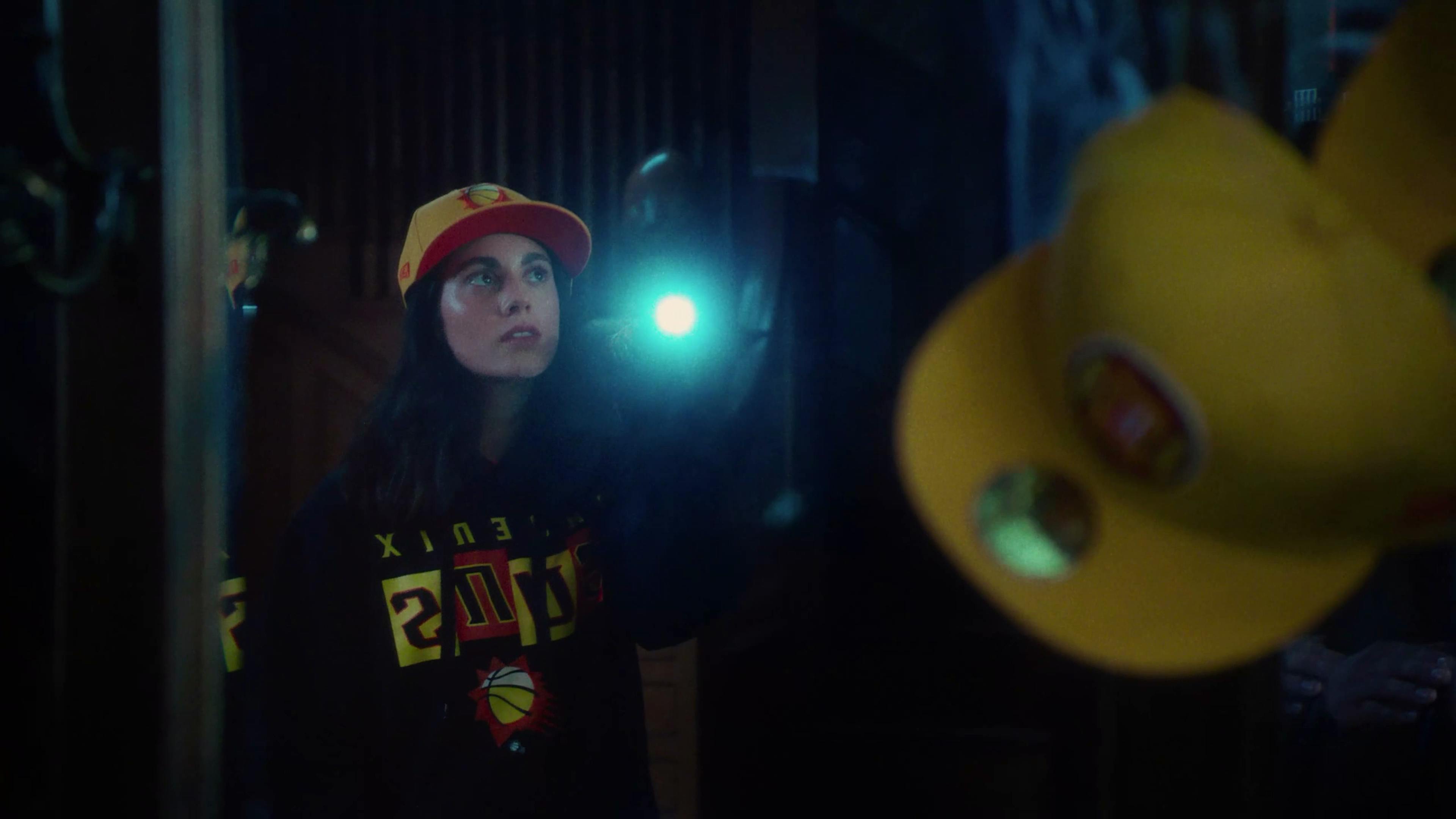 A person with a focused gaze, wearing a New Era cap and a basketball sweatshirt, holding a flashlight that illuminates their face in a dimly lit setting.