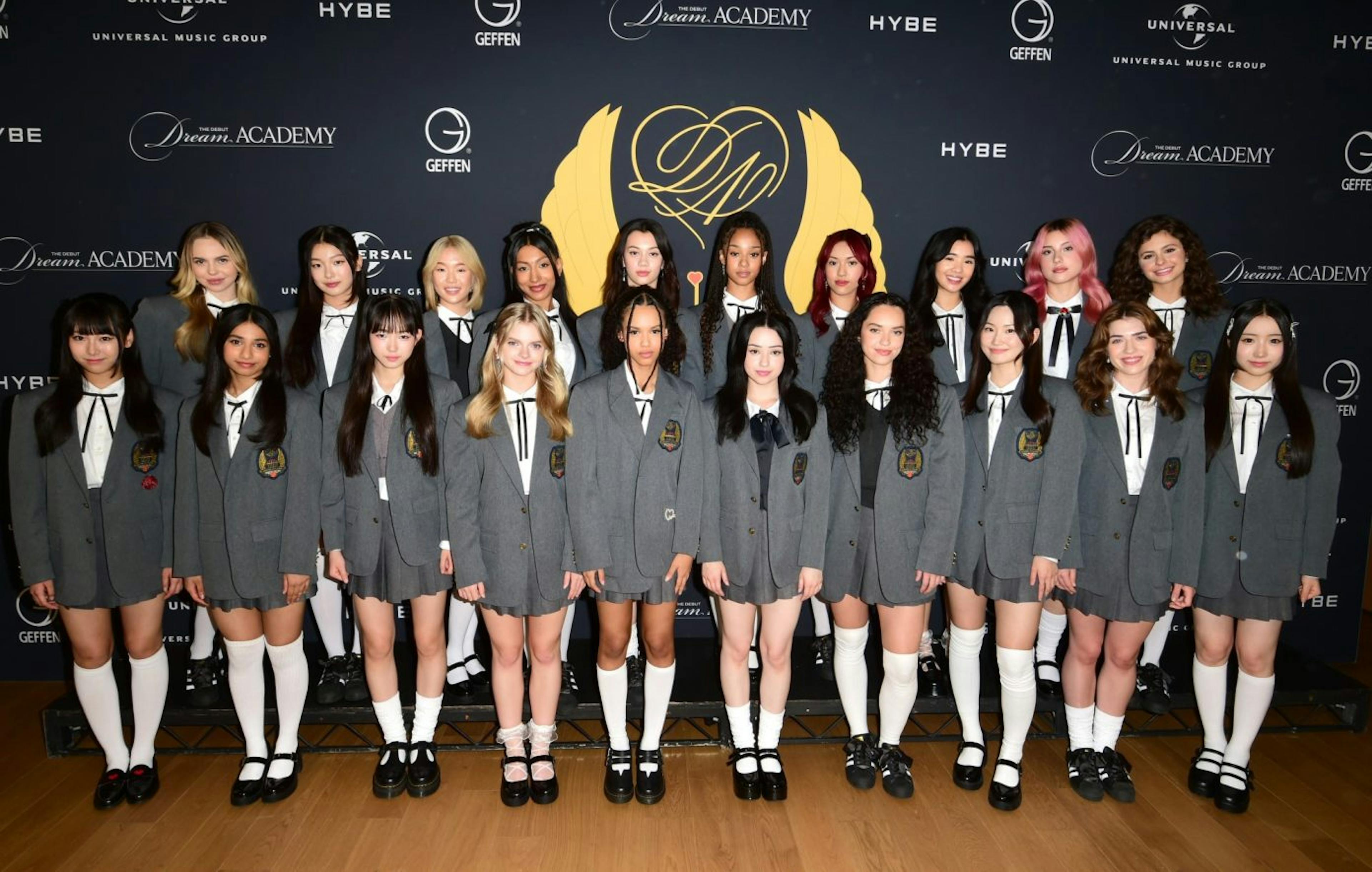 The image is a press photo for 'The Debut: Dream Academy.' It features a diverse group of young female contestants, all dressed in matching gray school-like uniforms with white shirts and ties, each with a unique badge symbolizing the academy. They stand in two rows, smiling and poised, in front of a backdrop that prominently displays the logos of HYBE, Geffen Records, and Universal Music Group, alongside the show's name. The contestants' varying hairstyles and confident stances reflect the global and inclusive nature of the talent competition, which aims to form an international girl group.