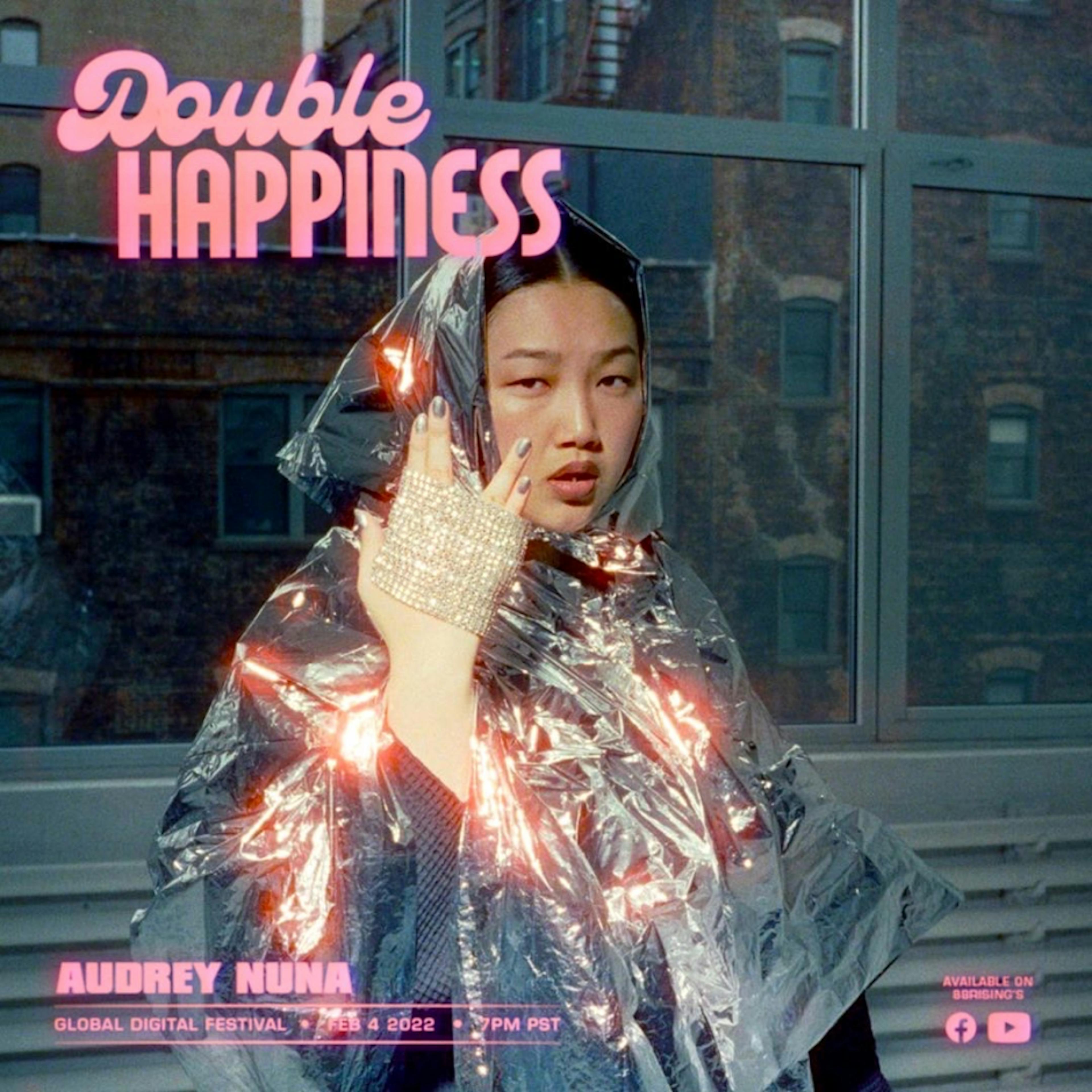 Promotional image for 'Double Happiness Global Digital Festival' featuring artist Audrey Nuna. She poses against an urban backdrop with apartment buildings visible through a window. Audrey is wearing a shiny, metallic rain poncho that catches the light, and her left hand is adorned with sparkling rings, raised to her face to draw attention. Her expression is cool and thoughtful. Above her, in neon-pink cursive, reads 'Double Happiness', and below in pink block letters are her name and the event details, noting the date as February 4, 2022, at 7 PM PST.