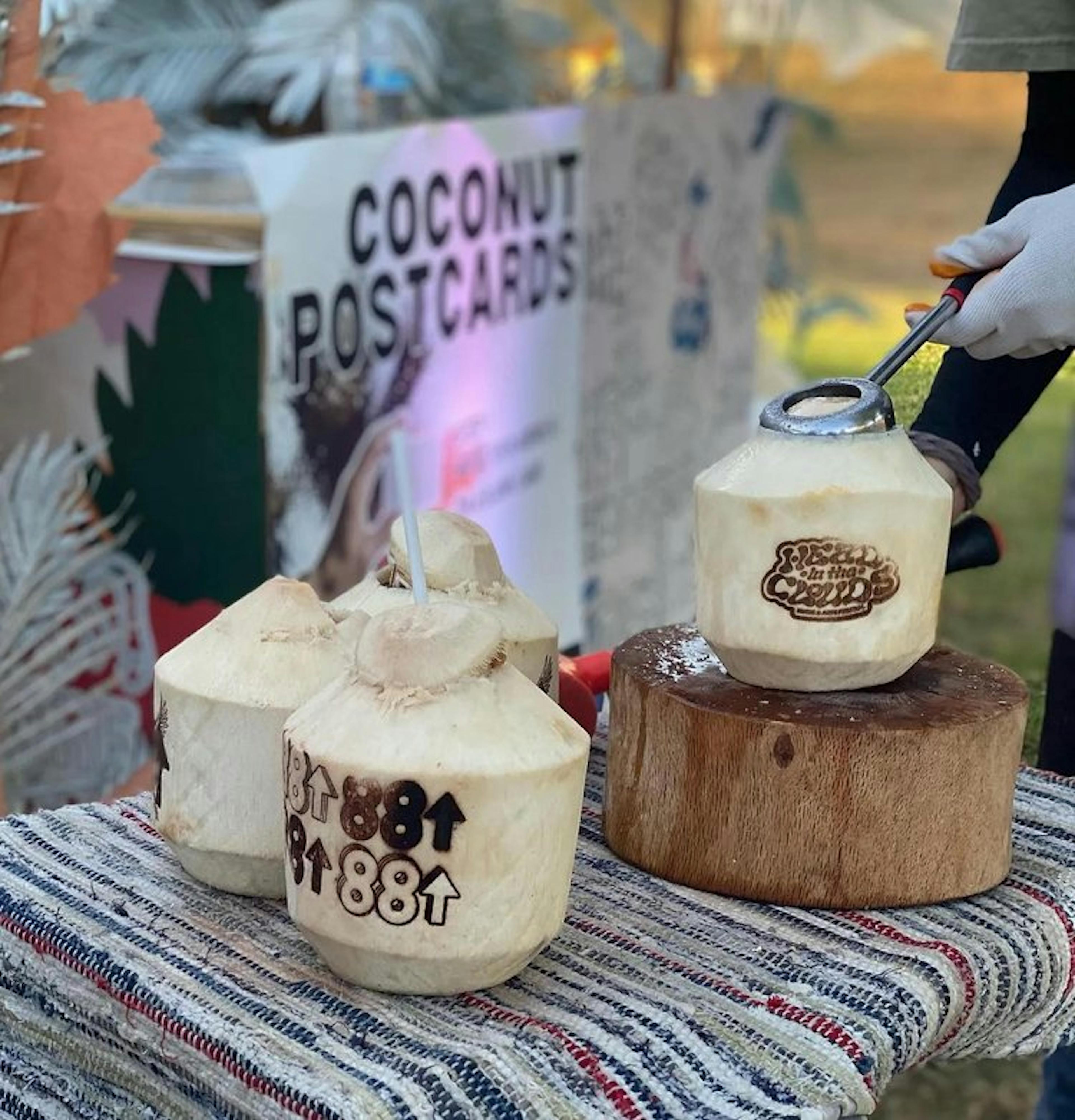 An inviting festival scene featuring fresh coconuts being prepared at the Head In The Clouds festival. Three coconuts are prominently displayed, each branded with the 88rising logo, and one is being opened with a tool by a person wearing white gloves. In the background, a sign reads 'Coconut Postcards,' suggesting an interactive festival experience. The setting is outdoors, with hints of a festival atmosphere suggested by the presence of various flags and decorations in soft focus behind the coconuts.