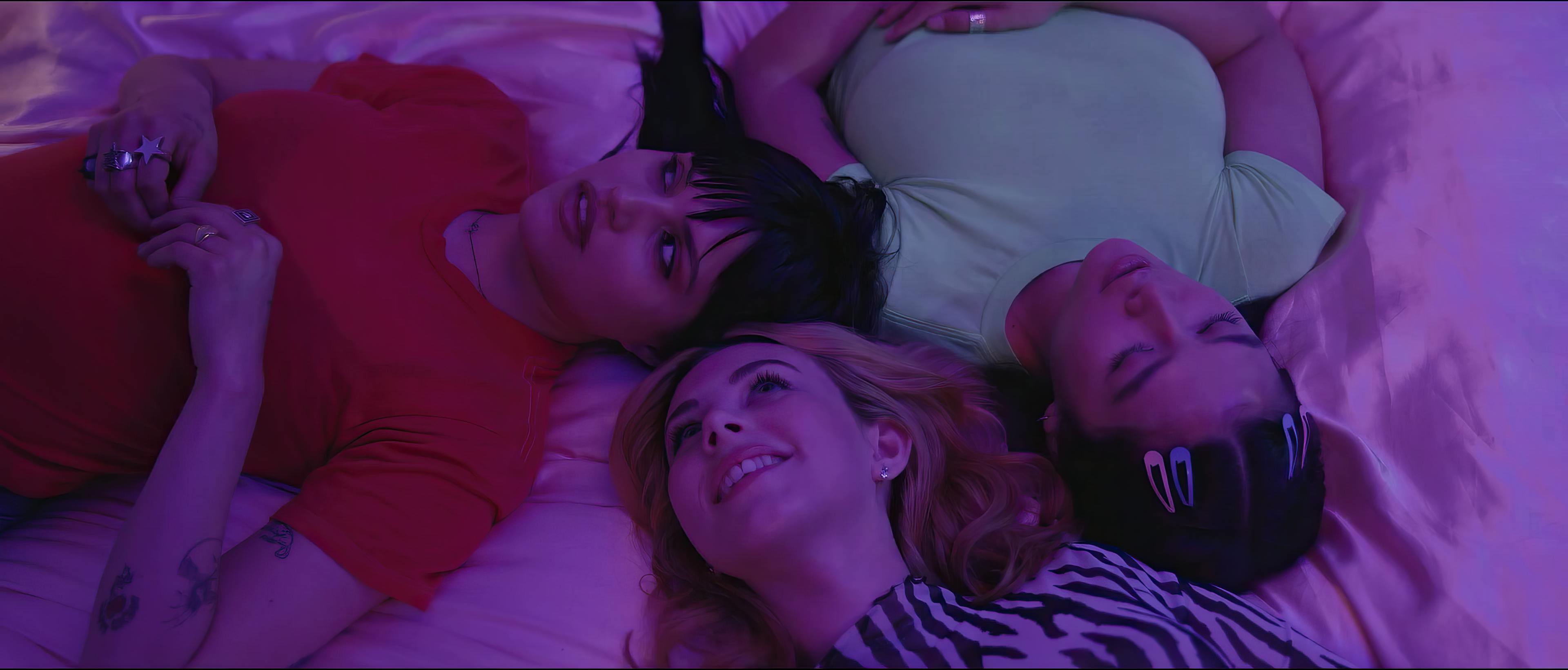 Three individuals lie close together on a bed, their heads forming a circle in the center. They appear relaxed and content in each other's company, illuminated by a soft, purple glow that sets a tranquil mood. The intimate arrangement and warm lighting suggest a scene of friendship and comfort.