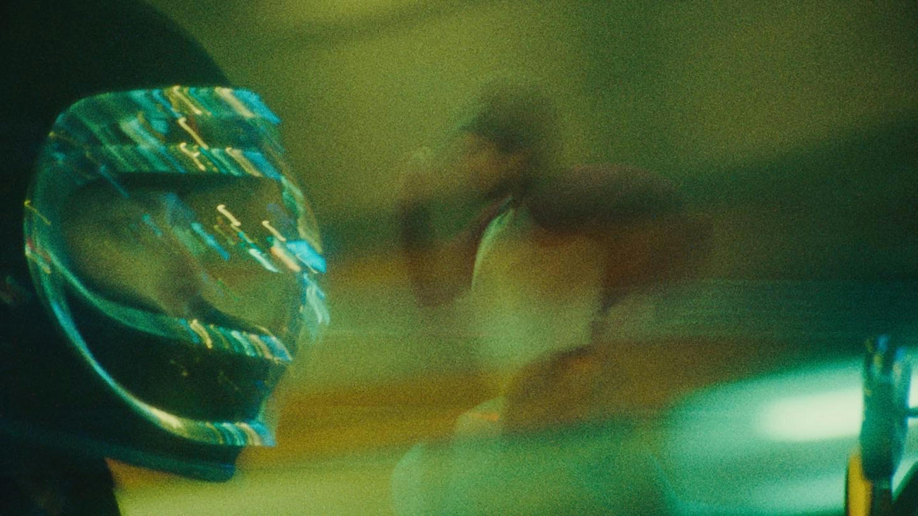 An abstract, motion-blurred photograph that captures a fleeting moment of action. In the foreground, a motorcycle helmet with a reflective blue visor is partially visible on the left, hinting at a sense of speed. To the right, the blurred silhouette of a person can be discerned, enveloped in a hazy, amber light that adds a mysterious quality to the image. The lighting and movement suggest a night scene, possibly on a city street, with the viewer getting just a glimpse of an urban adventure.