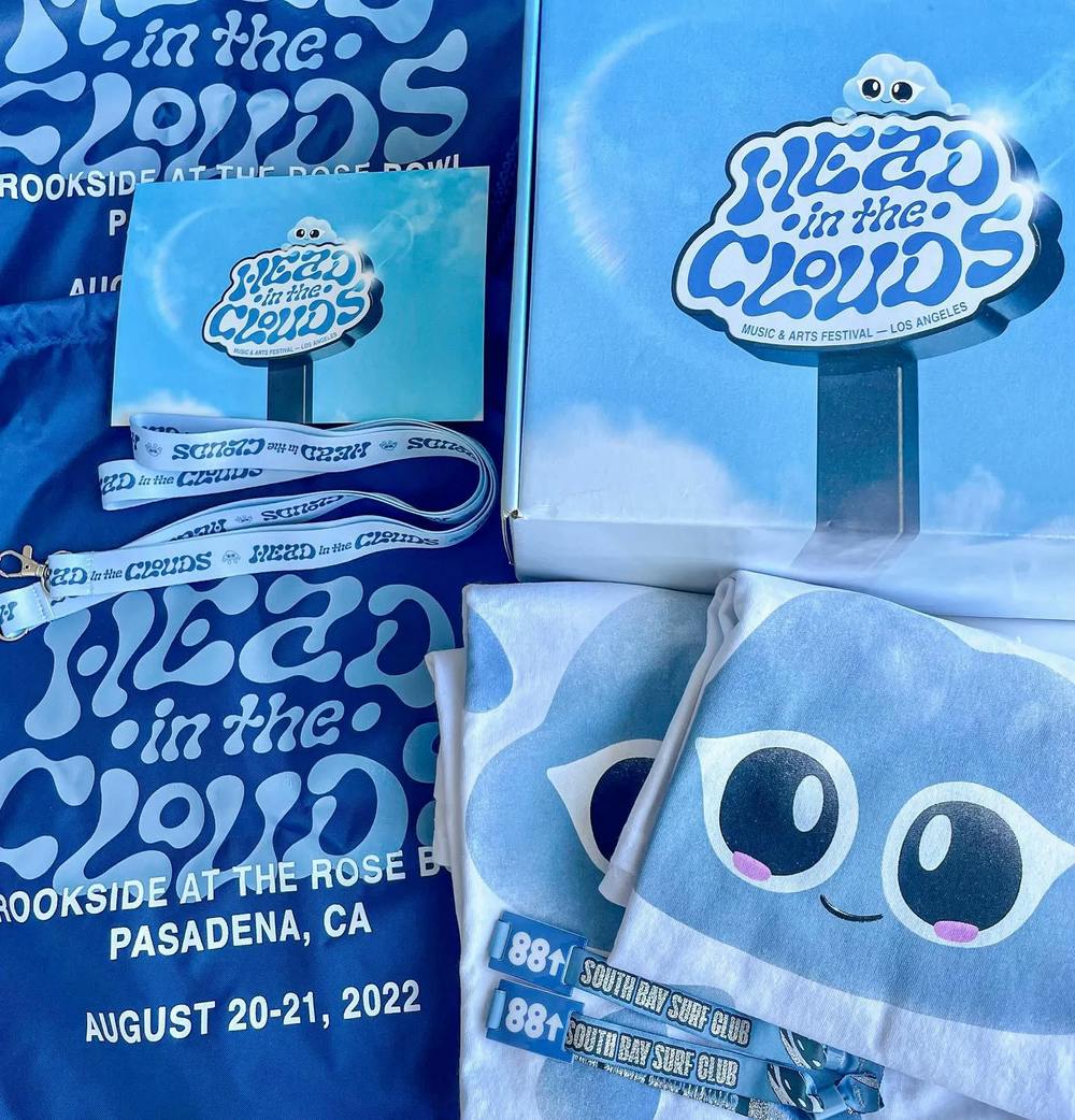 A collection of Head In The Clouds festival merchandise laid out on a blue background. Items include a light blue tote bag featuring Clo The Cloud God's face, a festival lanyard, a small square paper with the event's logo, and a folded blue t-shirt with the same branding. Each item carries the playful and friendly character of the festival's mascot and the cool blue tones of the event's aesthetic. 'Head in the Clouds' and the date of the festival, 'August 20-21, 2022', are prominently displayed, highlighting the event's details.