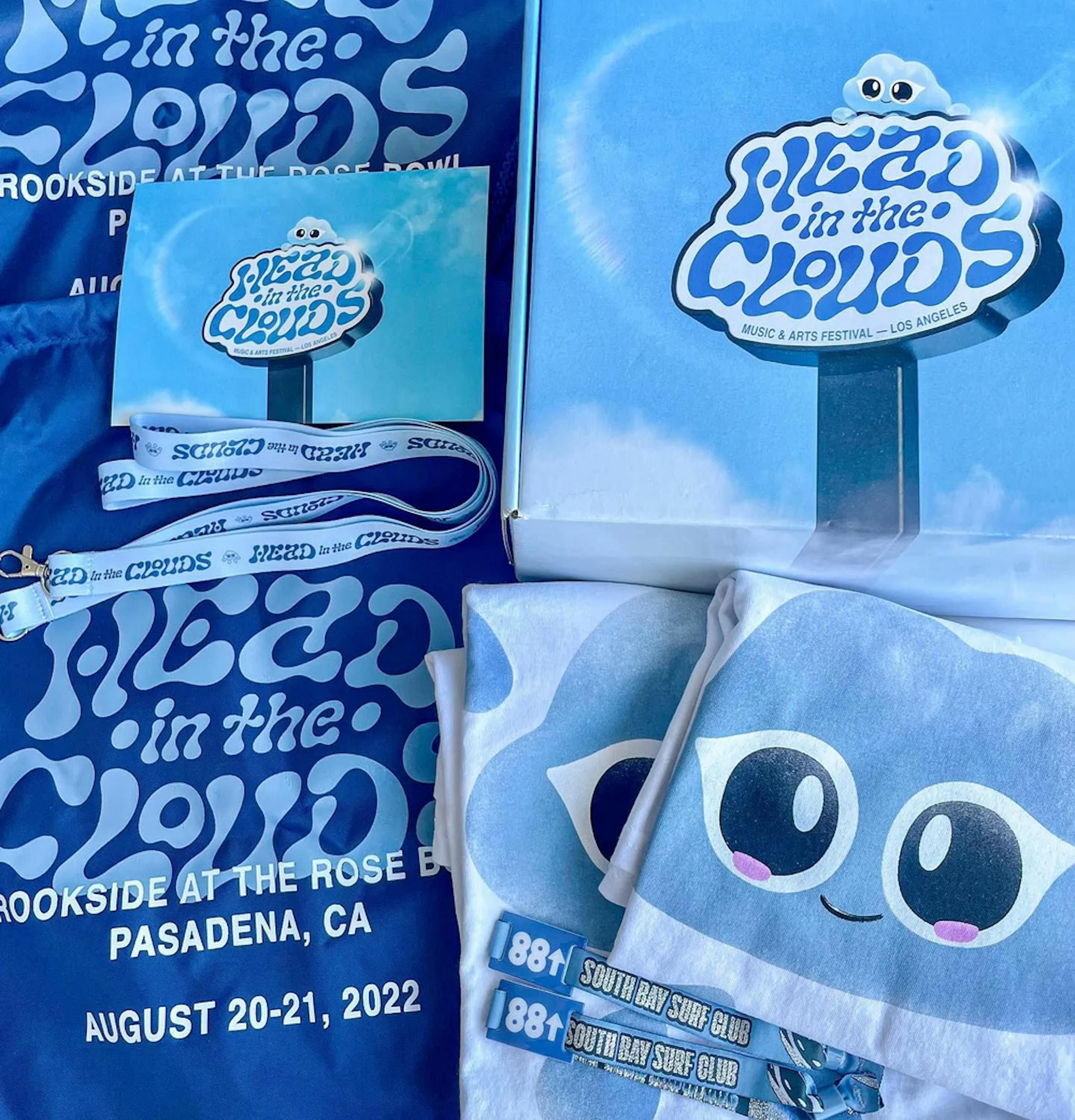 A collection of Head In The Clouds festival merchandise laid out on a blue background. Items include a light blue tote bag featuring Clo The Cloud God's face, a festival lanyard, a small square paper with the event's logo, and a folded blue t-shirt with the same branding. Each item carries the playful and friendly character of the festival's mascot and the cool blue tones of the event's aesthetic. 'Head in the Clouds' and the date of the festival, 'August 20-21, 2022', are prominently displayed, highlighting the event's details.