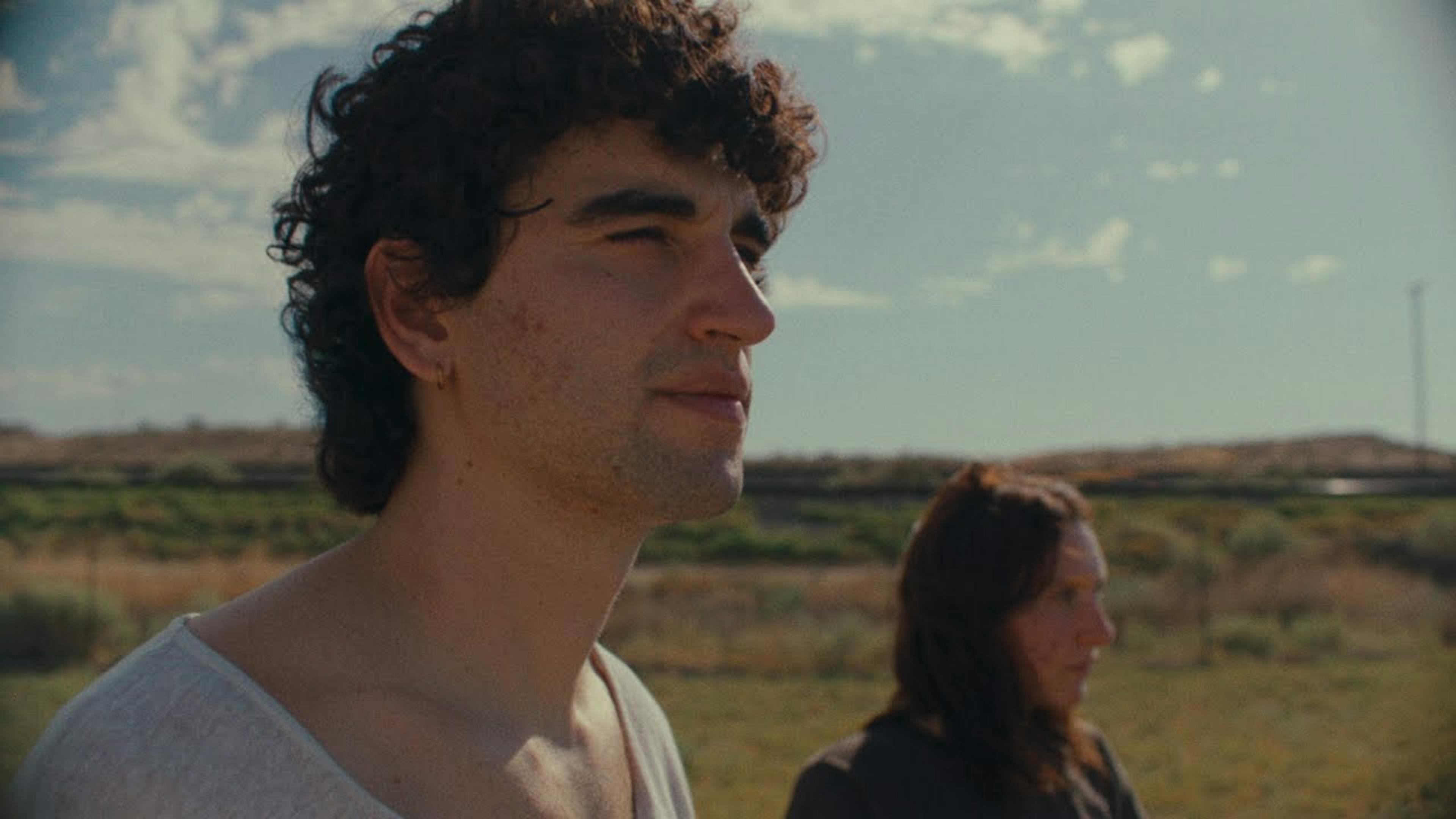 A still image from Carol Ades's music video 'Crying During Sex' captures a candid moment of two individuals outdoors. In the foreground, there's a man with curly dark hair, a slight stubble on his face, wearing a white, low-cut sleeveless top, gazing into the distance with a contemplative expression. Behind him and slightly out of focus, there is a woman with long, straight, brown hair, dressed in a black shirt, looking down thoughtfully. They are set against a serene backdrop of a clear blue sky and sparse vegetation in a dry, open field, evoking a sense of quiet introspection.