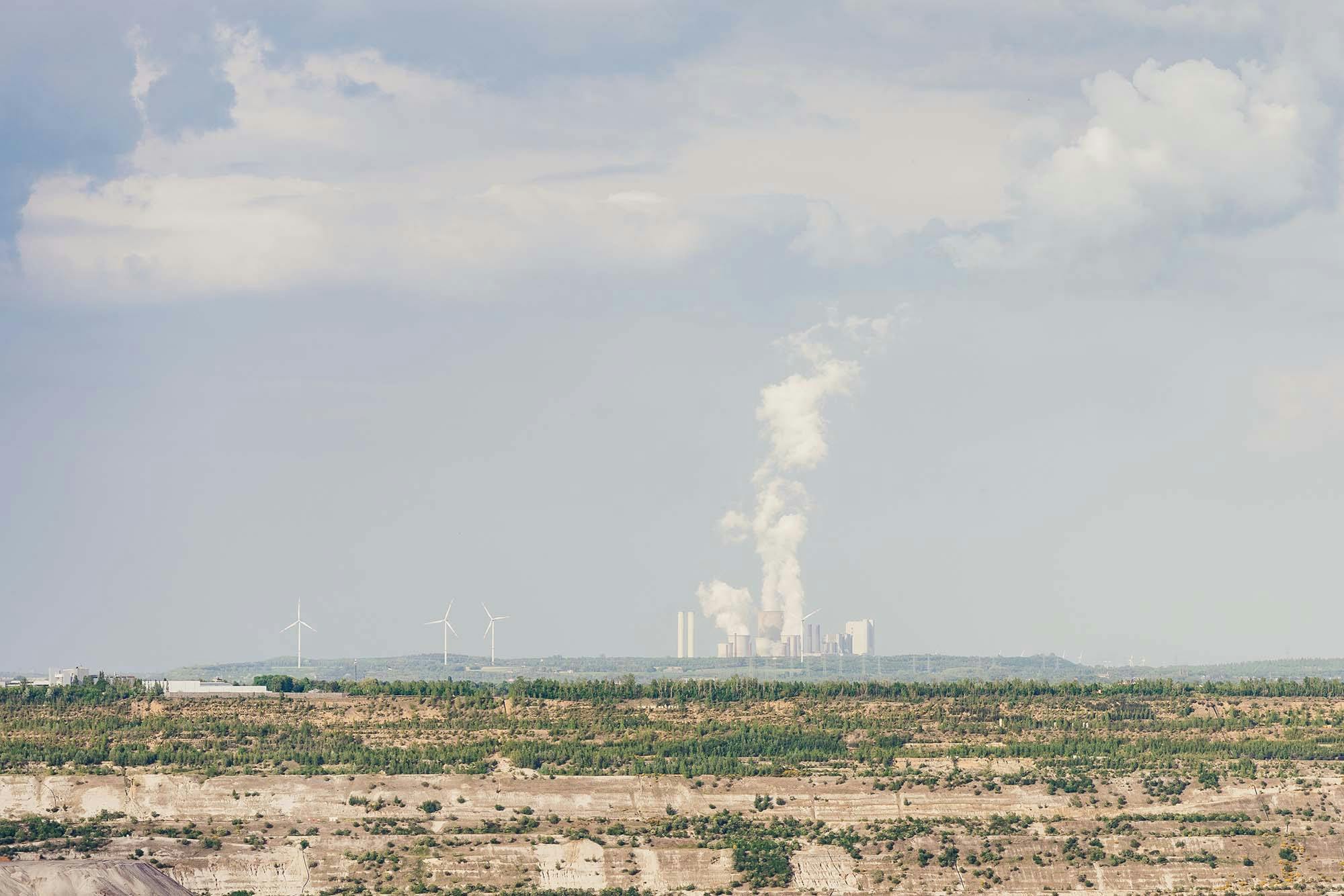 Smoke rises from factory chimneys (@barnimages). In front there are some wind mills and degraded land. 