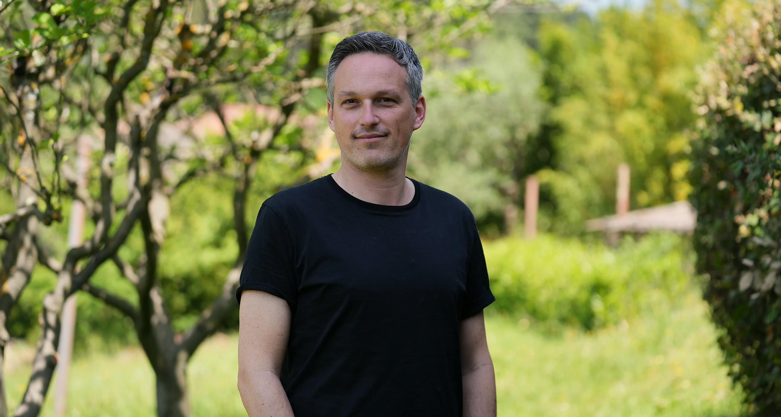 Jaron Pazi (male) standing in front of a park with trees and bushes.