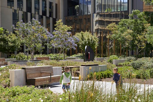 A photograph of two children playing in large public courtyard full of plants. There are various seating areas, with trees and large building facades in the background.