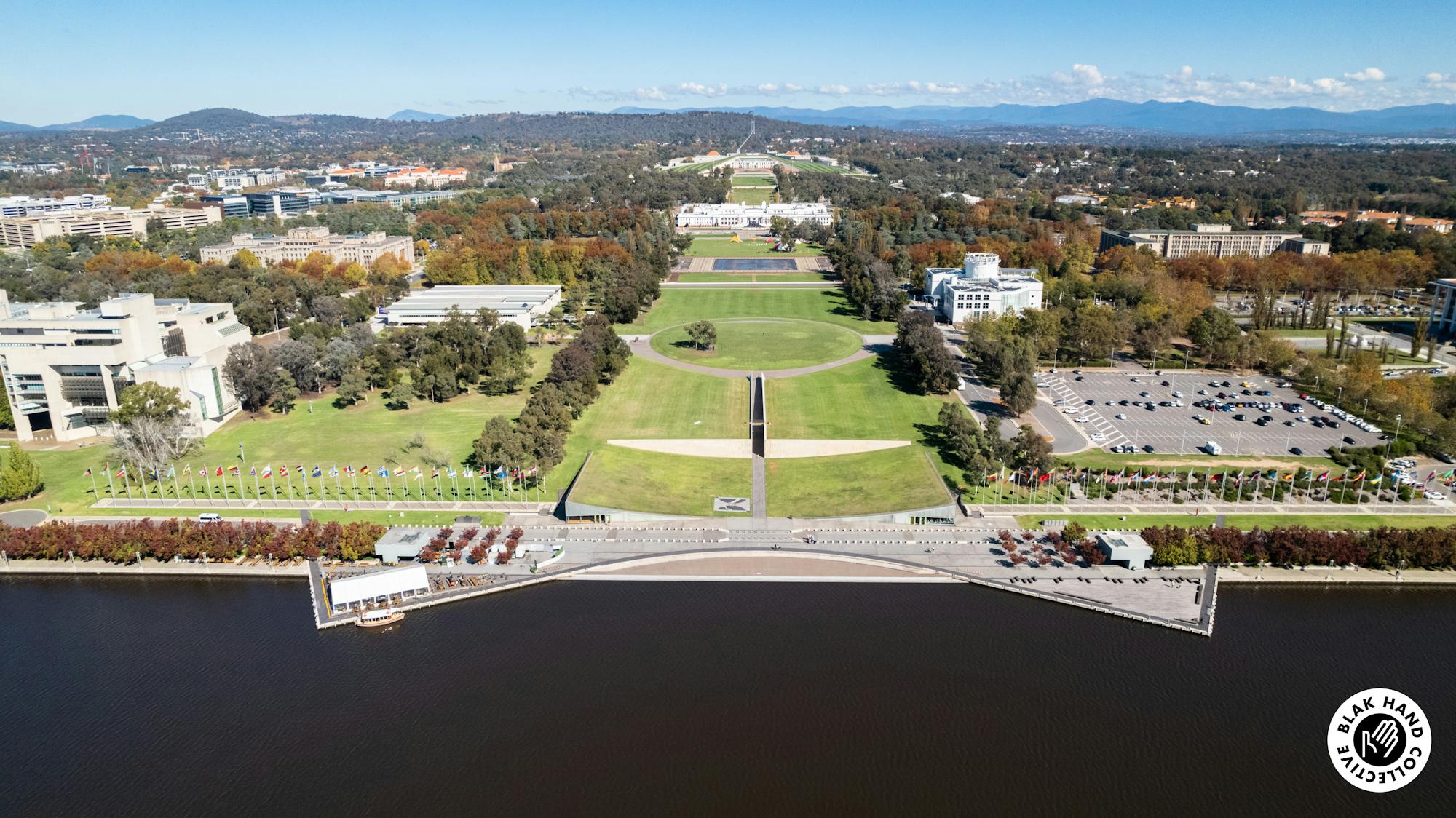 An aerial photograph of a lake front site. There is an large open corridor of grass running though the middle of the image, trees and buildings surround the open area, with mountains and blue sky visible at the top of the image. 