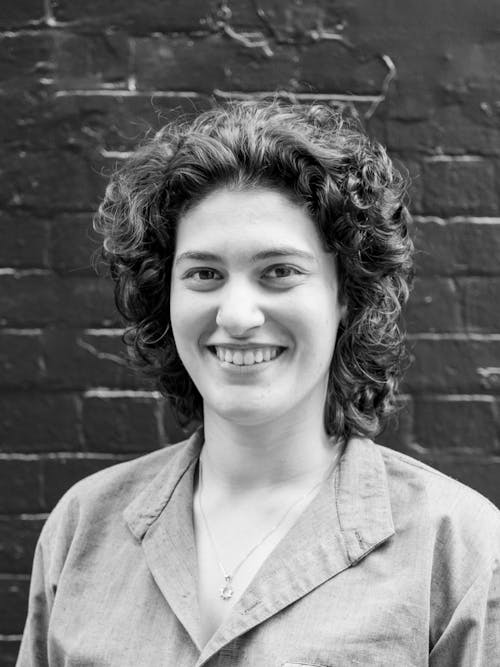 A black and white headshot of a person with curly hair in front of a brick wall painted black