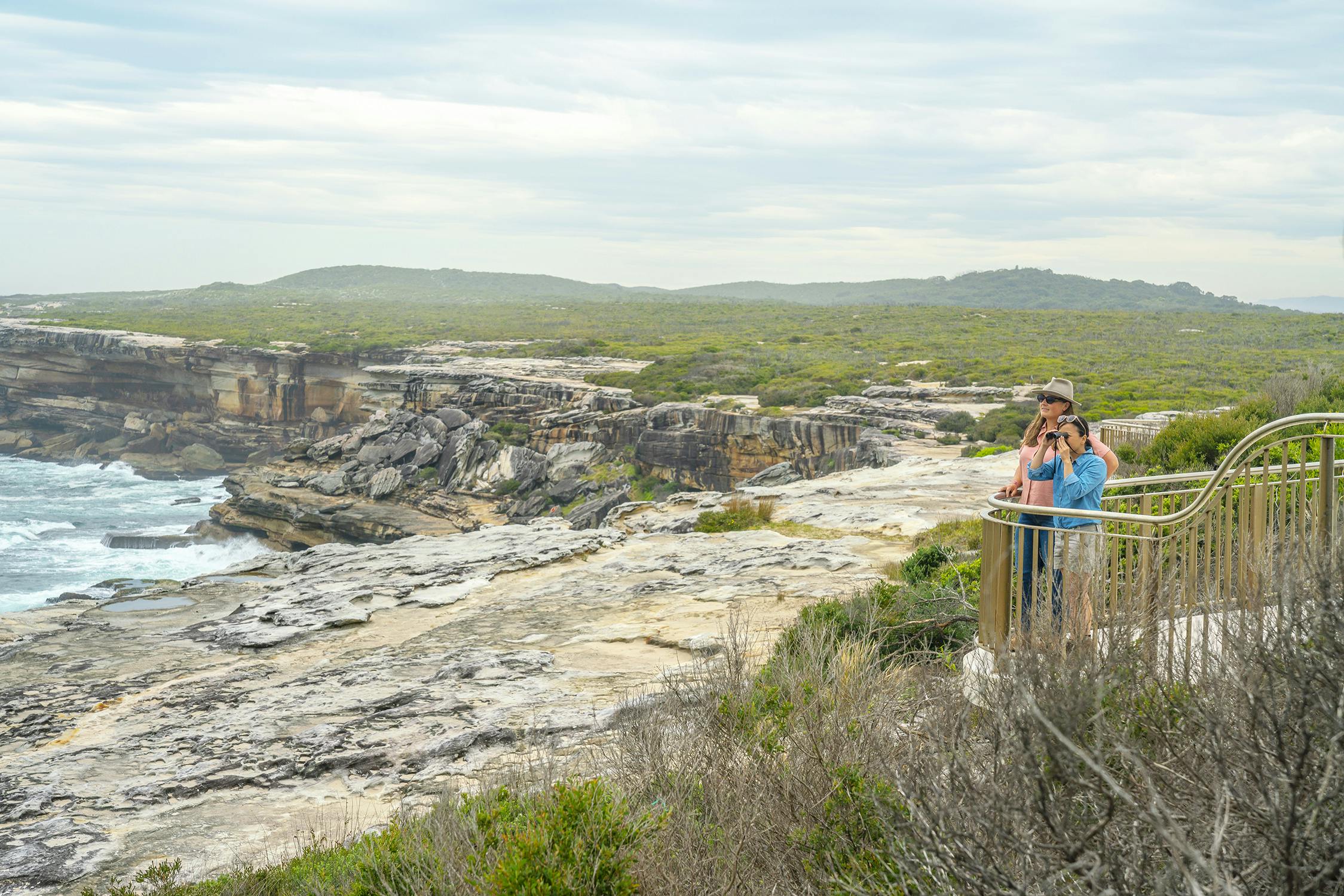 A photograph of two people standing on a viewing platform overlooking a dramatic sandstone coastline. One is wearing a hat, the other is looking through binoculars. There is vegetation in the foreground and into the distance.