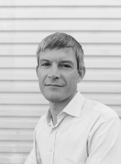 A black and white headshot of a person wearing a white shirt in front of a white wall.