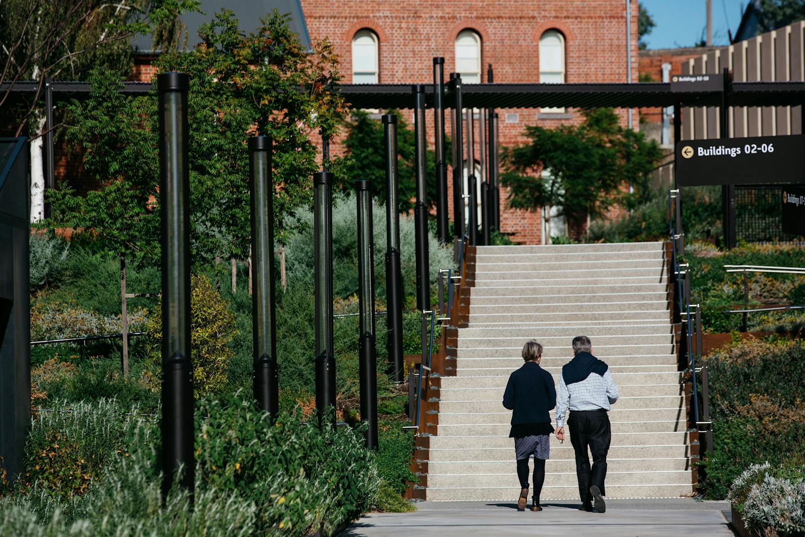 A photograph of two people walking towards an outdoor staircase, with plants either side. There is a red brick building at the top of the stairs, and black light fixtures to the left of their path.