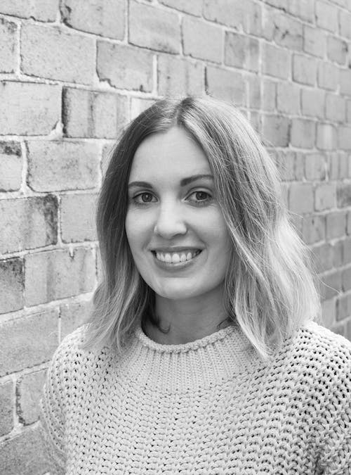 A black and white headshot of a smiling person in front of a brick wall.