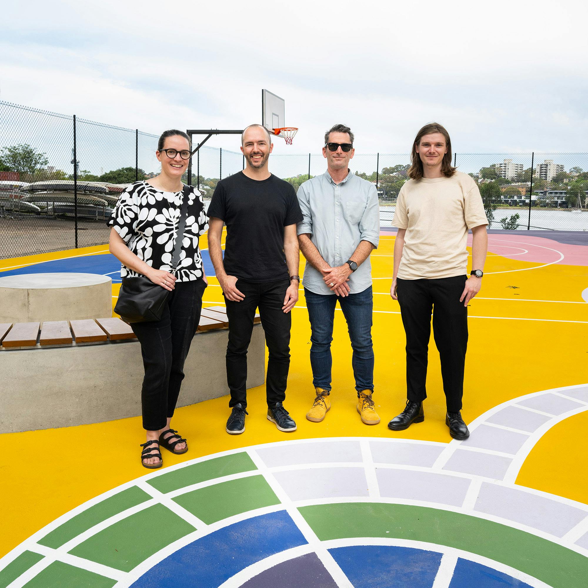 Four people posing for a photo outside on a brightly painted ground. There is a curved bench beside them and a basketball hoop in the background.