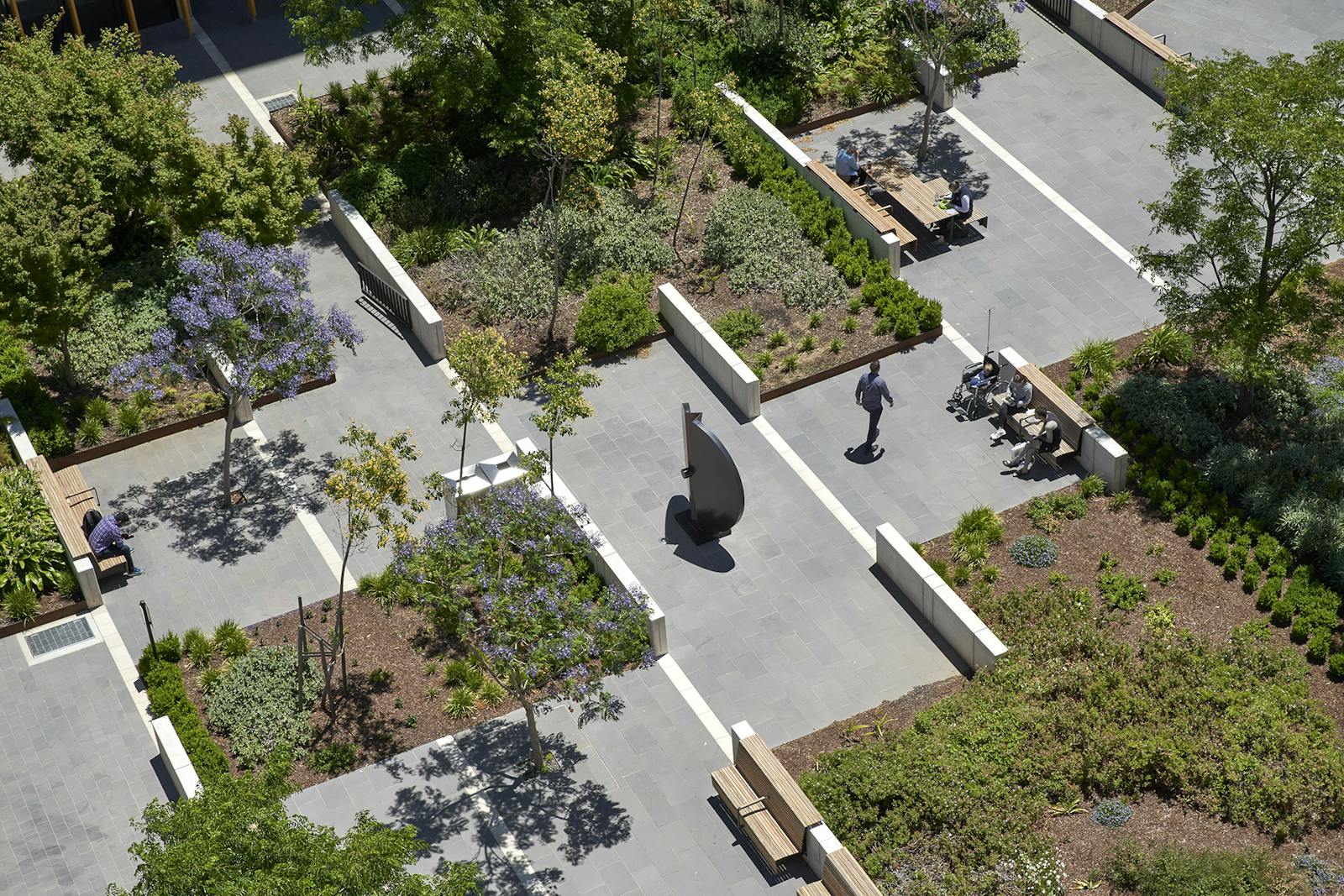A photograph a person walking through large public courtyard. It's a patchwork of grey paved and green planted areas.