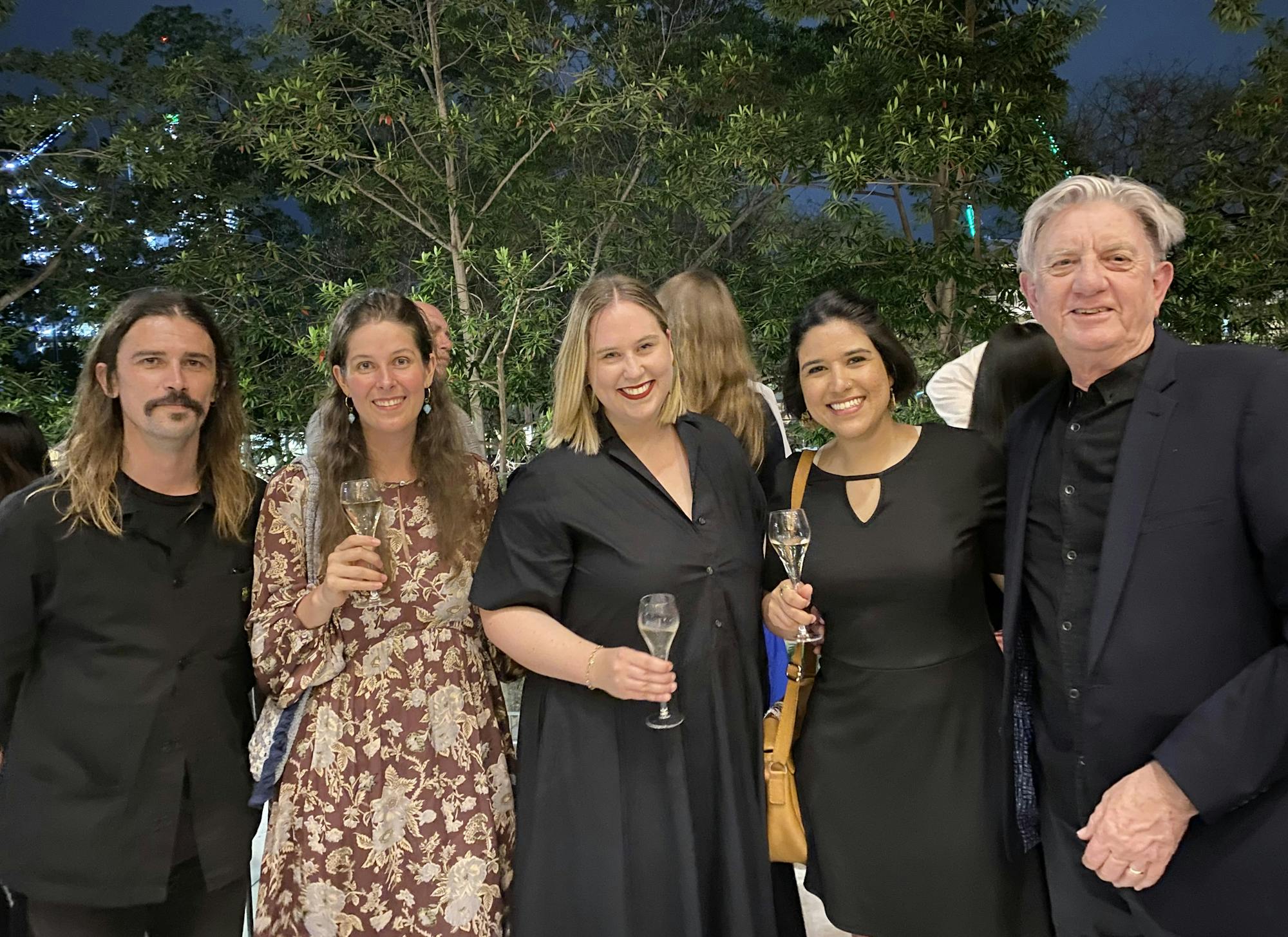 A photograph of five people in a row, smiling at the camera. Four are wearing dark colours, three are holding wine glasses, two have short hair. Above their heads we can see trees and a night blue sky.