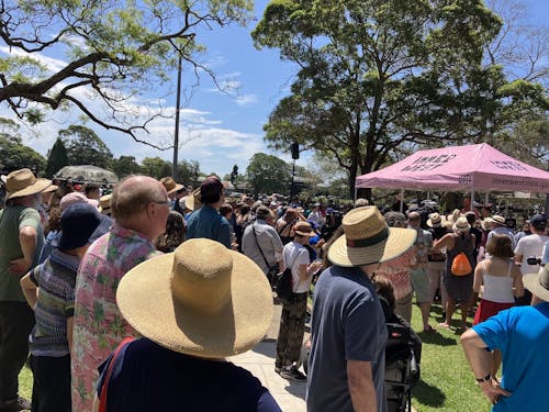A photograph of a large group of people gathering in a sunny park, everyone is facing towards a tent with a pink roof, listening to a speech. Trees and blue sky is visible in the background.