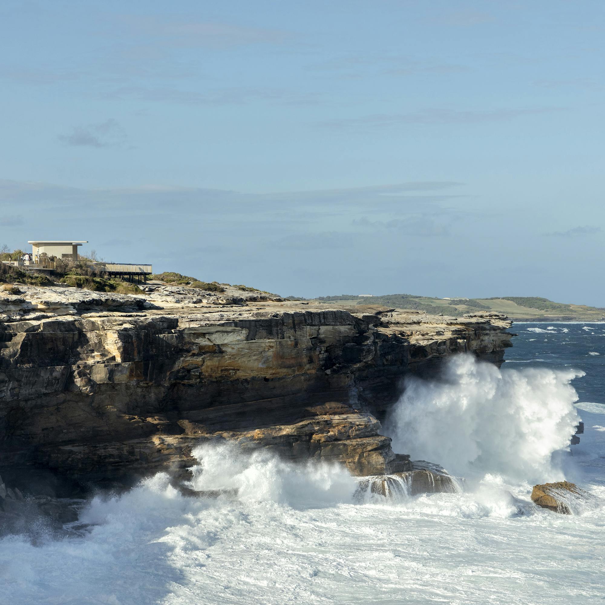 A photograph of large ocean waves crashing against a sandstone cliff, the sky is blue with scattered clouds, and there is a viewing platform blending into the landscape on the top of the cliff.