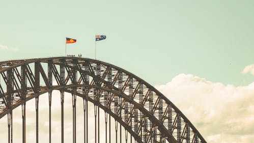A photograph of the top of a steel arched bridge (The Sydney Harbour Bridge) with Aboriginal and Australian flags flying at full mast. There are a few people seen in silhouette near the top. 