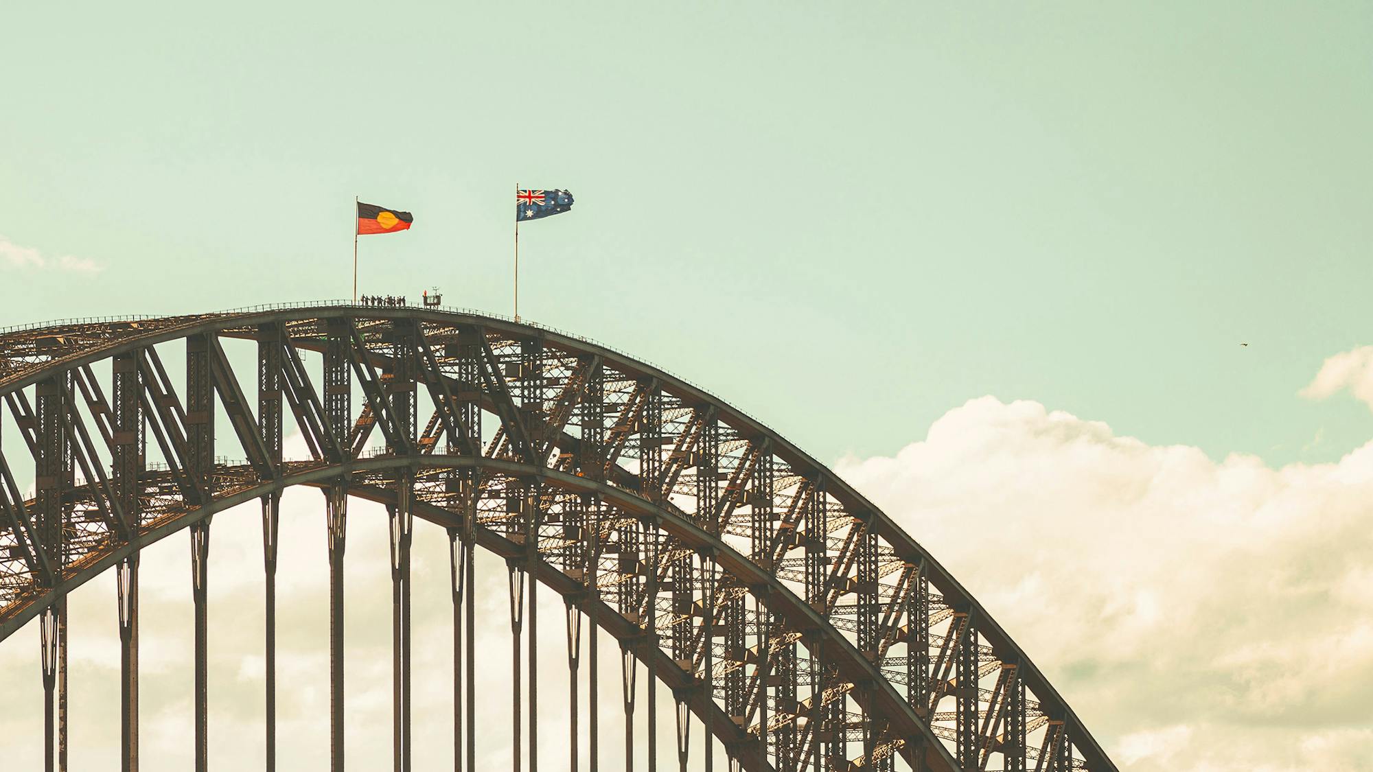A photograph of the top of a steel arched bridge (The Sydney Harbour Bridge) with Aboriginal and Australian flags flying at full mast. There are a few people seen in silhouette near the top. 