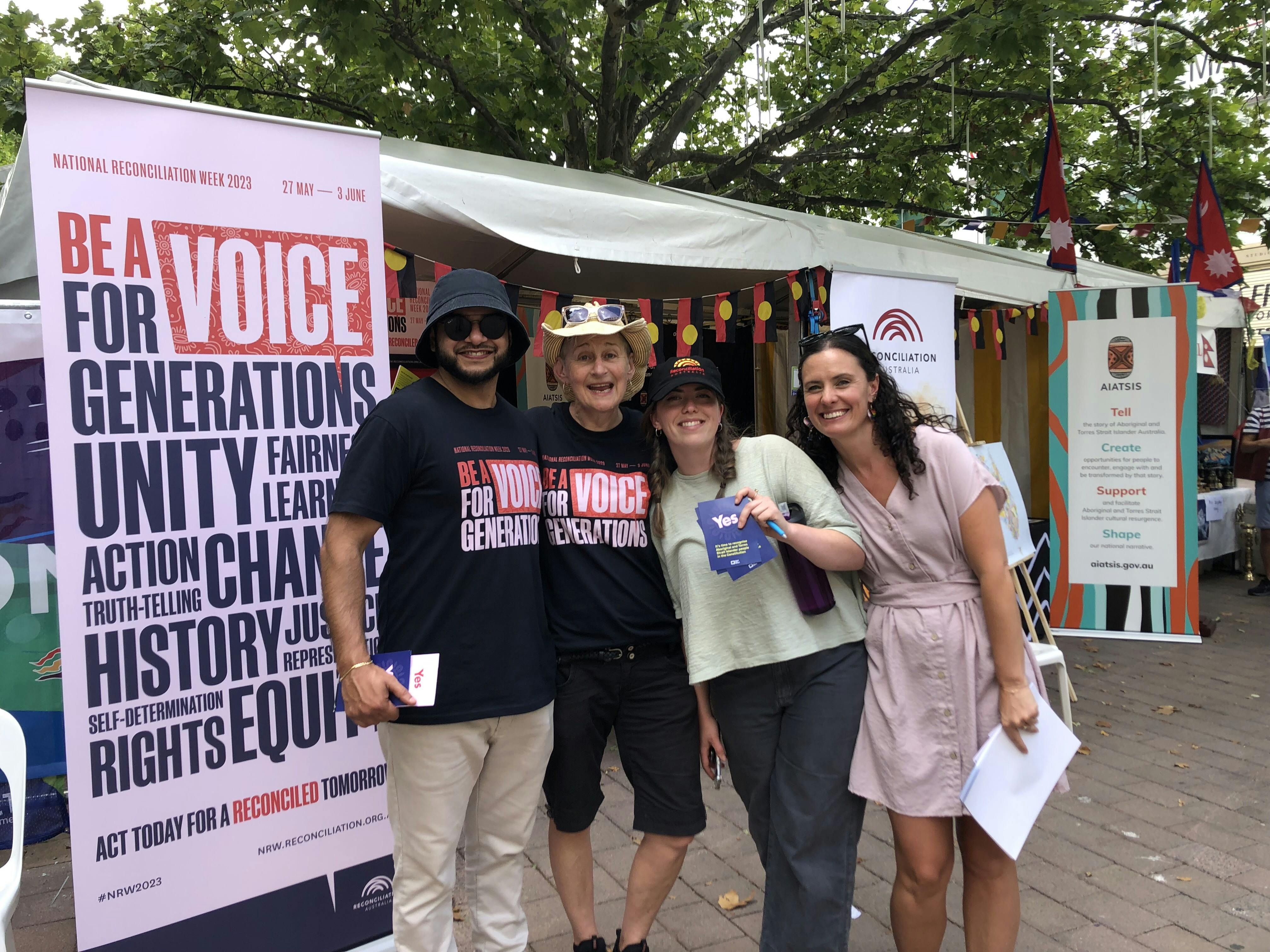 A photograph of four people smiling at the camera in front of a market stall. Two of the people are wearing t-shirts which say 'be a voice for generations'