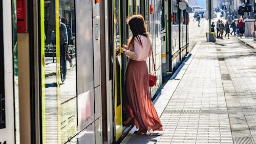 A photograph of person in a long flowing skirt stepping onto a tram in urban Melbourne. There are other people in the distance, and a reflection of a cyclist in the tram doors.