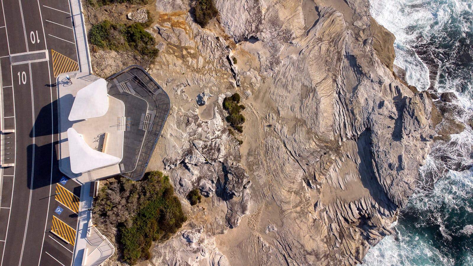 An aerial photograph of a sandstone coastline. On the left a contemporary white viewing platform and road is visible. On the right of the image, waves crash against cliffs.