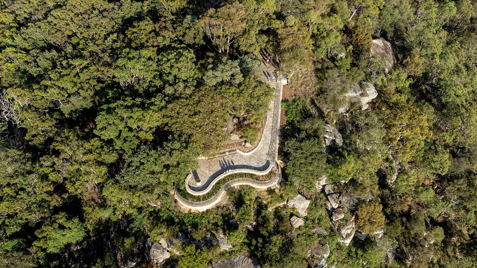 An aerial photograph of a curvy, sandstone lookout surrounded by green bushland, with people arriving and sitting on a low wall.