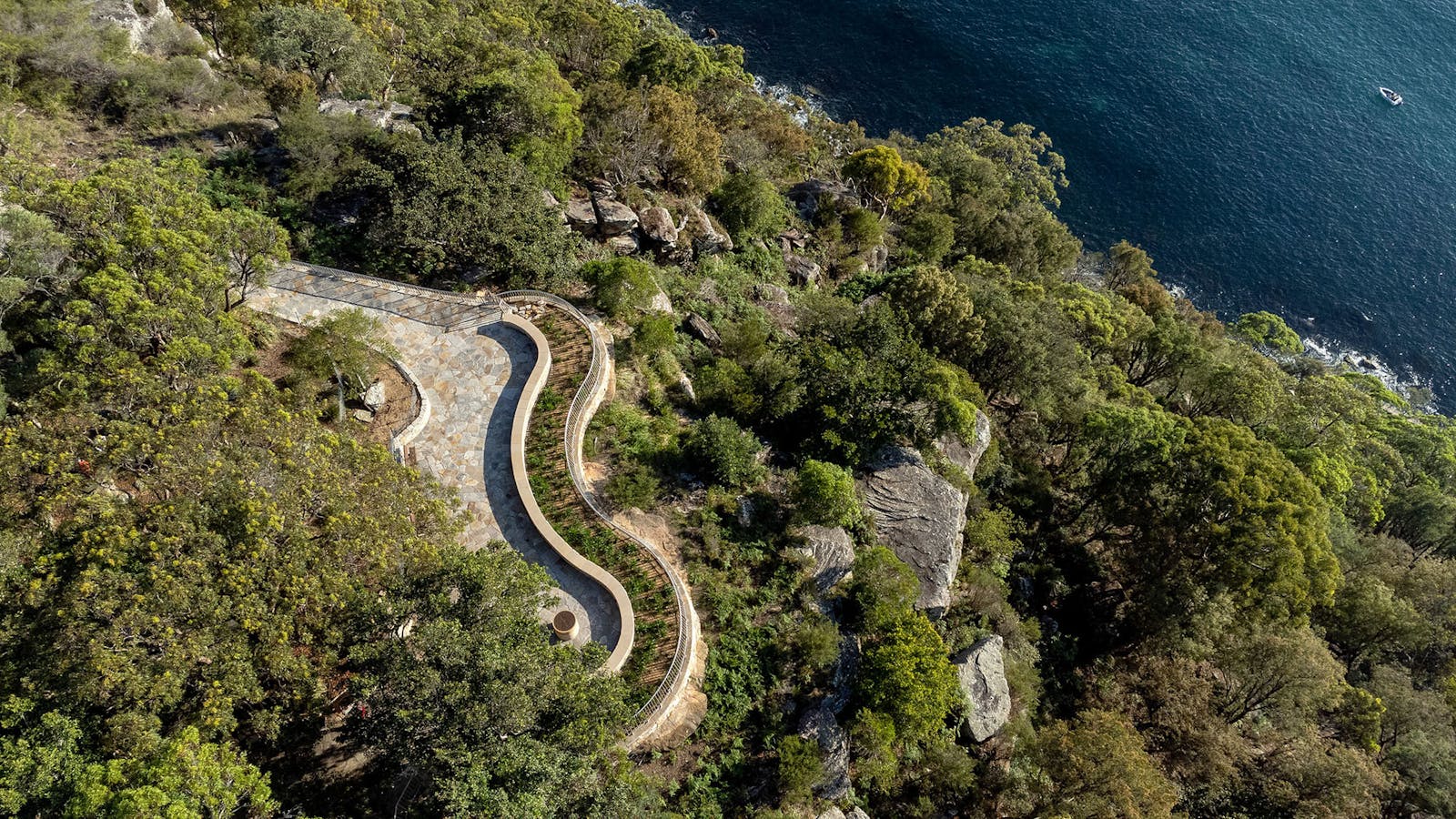 An aerial photograph of a curvy, sandstone lookout surrounded by green bushland, with water and a boat below.
