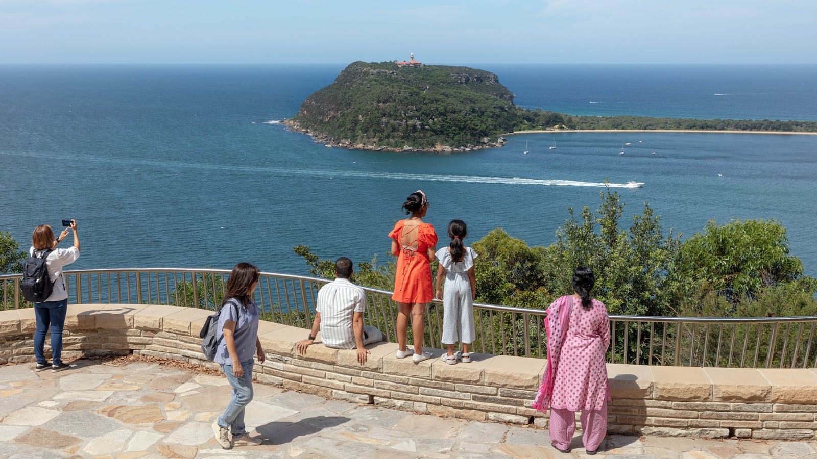 A photograph of people standing, sitting, photographing, and walking, at a Sydney sandstone lookout with water views.