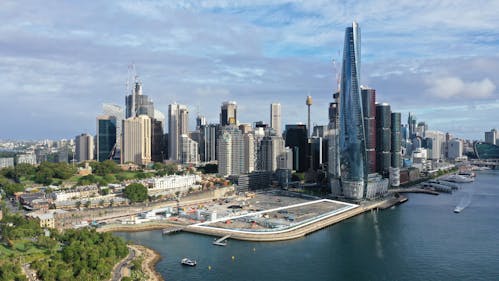 An aerial photograph of Sydney, with the harbour and Barangaroo Precinct in the foreground, and the city and a cloudy sky behind.  A white outline has been added to indicate the Harbour Park site perimeter, as part of a larger construction site.