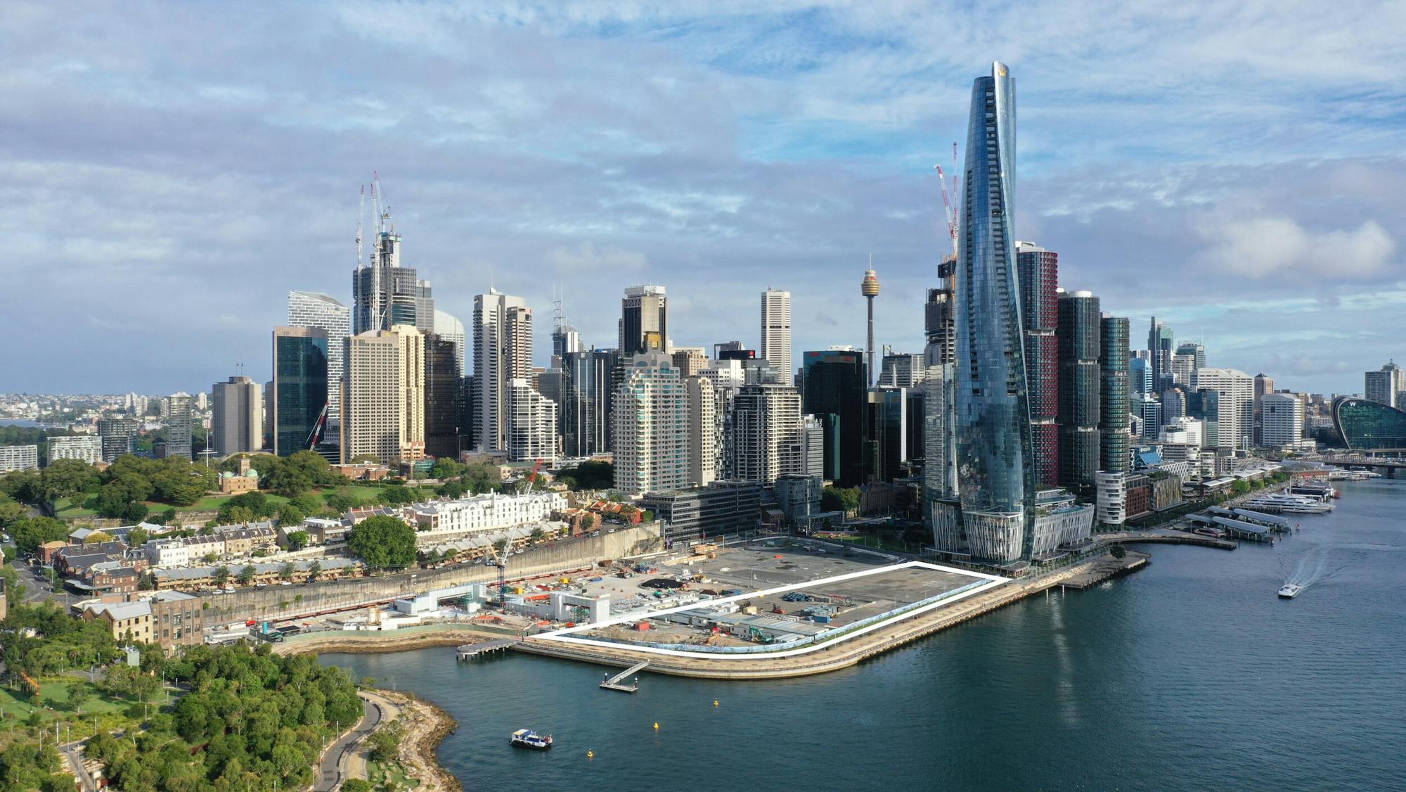 An aerial photograph of Sydney, with the harbour and Barangaroo Precinct in the foreground, and the city and a cloudy sky behind.  A white outline has been added to indicate the Harbour Park site perimeter, as part of a larger construction site.